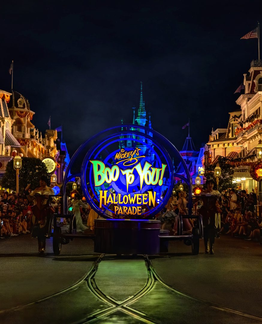 A large illuminated round marquee in a glowing blue frame, reading Mickey's Boo-To-You Halloween Parade in green and gold lettering, mounted on a parade unit rolling down Main Street at night, with the spires of Cinderella Castle lit green and blue behind it and crowds along both sides