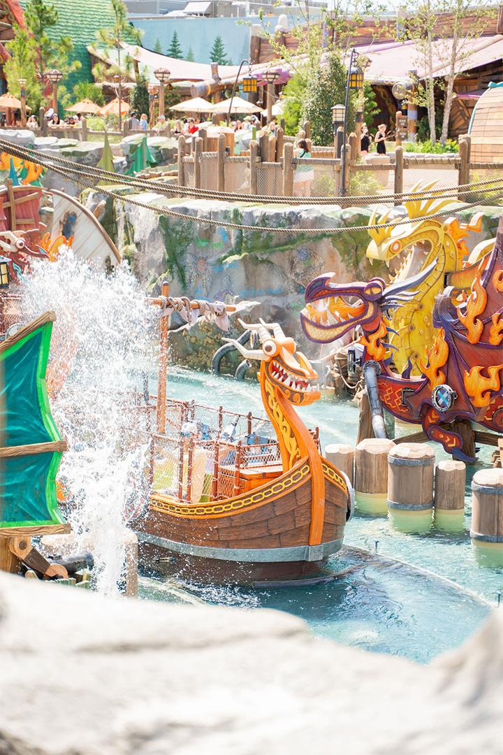 A wooden Viking longboat with an orange dragon-head prow floating in a turquoise splash pool, water spraying up beside it, with a large purple and gold dragon figurehead and rustic Isle of Berk buildings behind