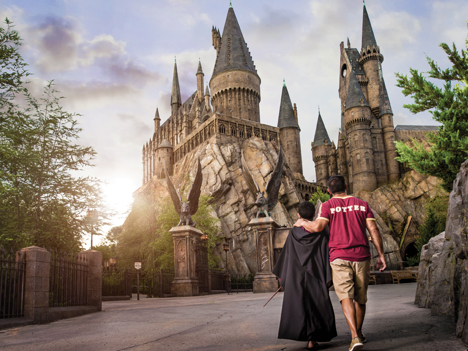 Two guests seen from behind, one in a black Hogwarts robe carrying a wand and a taller companion in a maroon Potter jersey with an arm around their shoulders, walking up the path past the winged boar statues toward Hogwarts Castle in low golden light