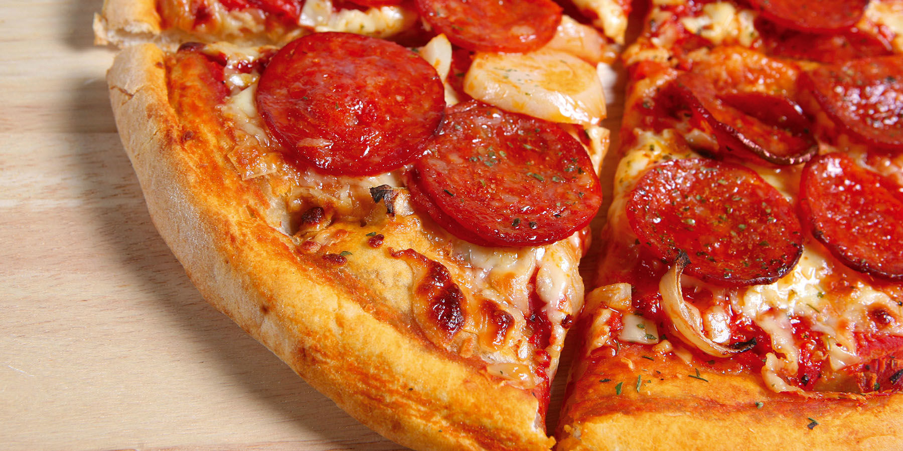 Close up of a sliced pepperoni and onion pizza on a light wood surface with a puffy golden crust, curled browned pepperoni and melted mozzarella