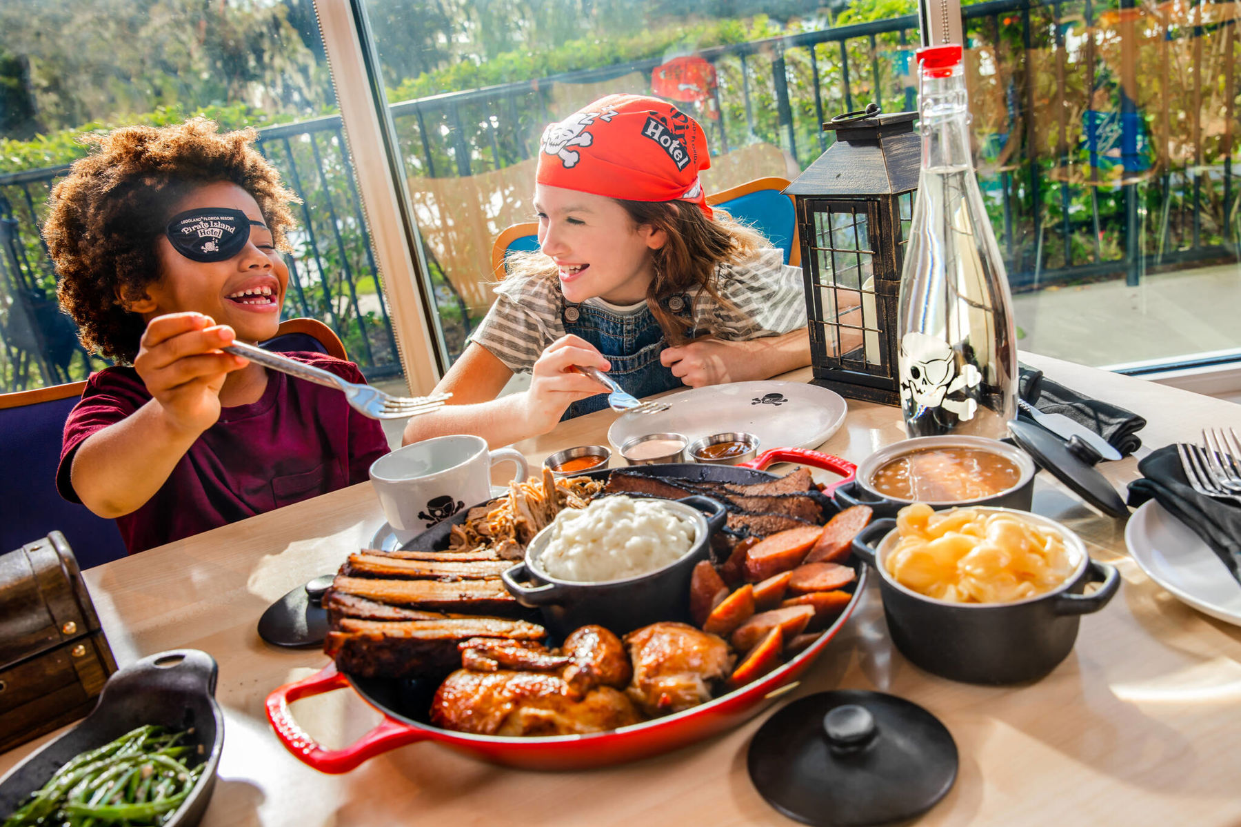 Two children in pirate eyepatch and bandana laughing at a sunlit table at Shipwreck Restaurant behind a large cast iron platter of ribs, brisket, pulled pork, roast chicken and sausage with crocks of mashed potato, gravy and macaroni cheese, with green beans alongside