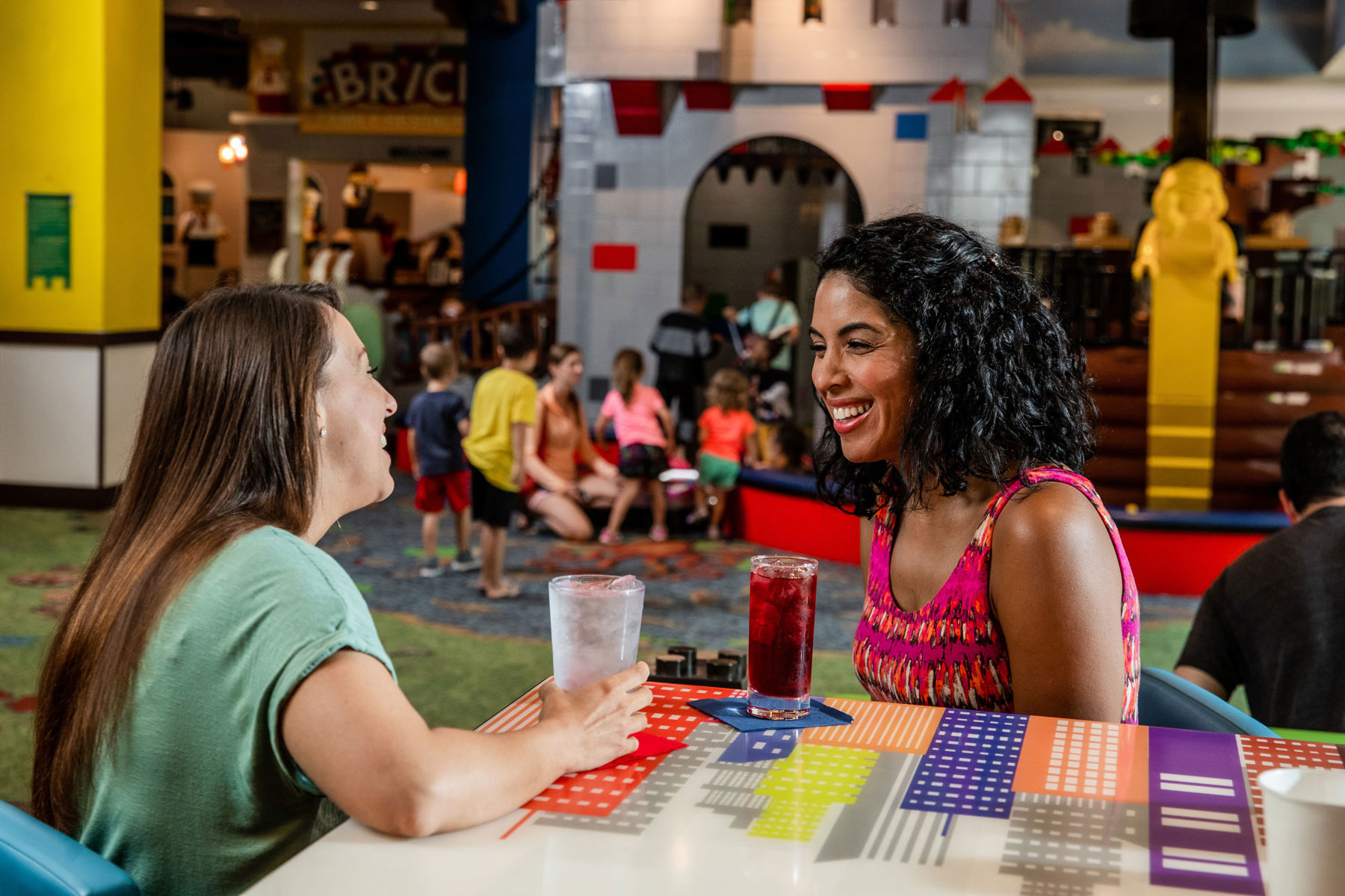 Two women laughing across a table with a city skyline print top inside LEGOLAND Hotel, with a BRICKS sign, a LEGO brick castle play structure, a giant yellow minifigure column and children playing on LEGO print carpet behind them