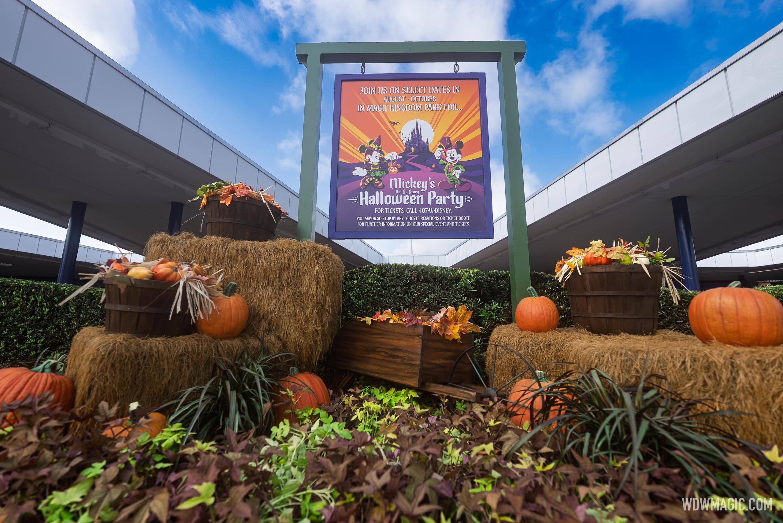 A hanging purple and orange Mickey's Not-So-Scary Halloween Party sign in a green wooden frame, surrounded by hay bales, pumpkins and bushel baskets of gourds, with elevated walkway ramps rising overhead on both sides