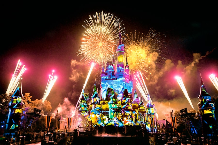 Cinderella Castle covered in blue, green and yellow projections at night, with gold and pink fireworks bursting overhead and streaming down either side