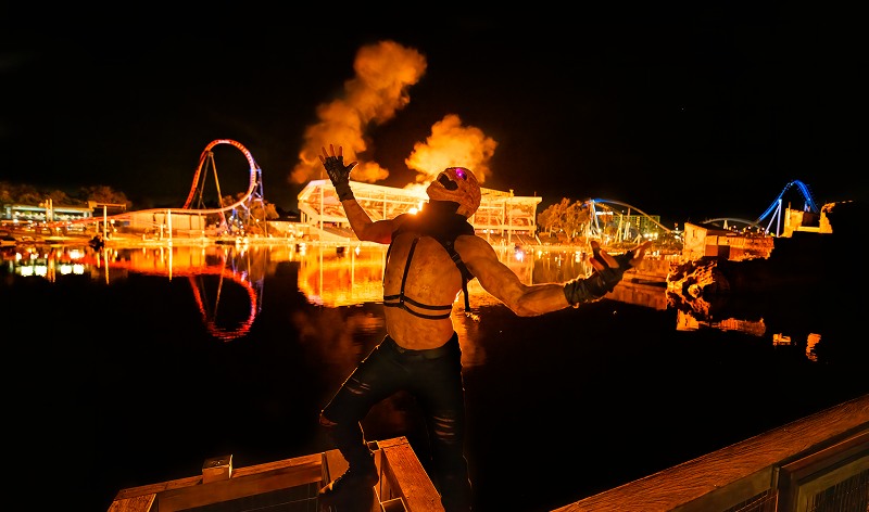 A costumed scare actor in a pale full-head mask stands among wooden dock timbers at night, arms outstretched and lit orange, as two large fireballs erupt over the water behind him with a brightly lit coaster loop on the far shore