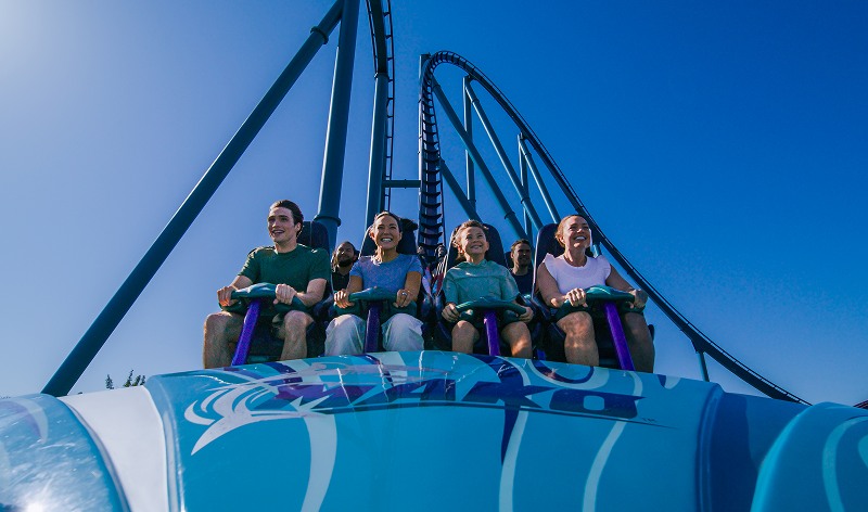 Front-row riders on a Mako train against a clear blue sky, gripping teal handles on purple restraints, with the Mako wordmark across the blue nose cone and the coaster track curving up behind them