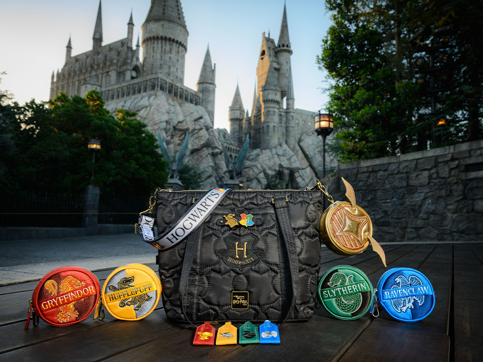 A black quilted Hogwarts tote with a white HOGWARTS strap and two charms clipped to its front tab, flanked by four round house coin pouches and a round Golden Snitch coin pouch, with four house zipper pulls lined up in front, on a boardwalk below Hogwarts Castle at dusk