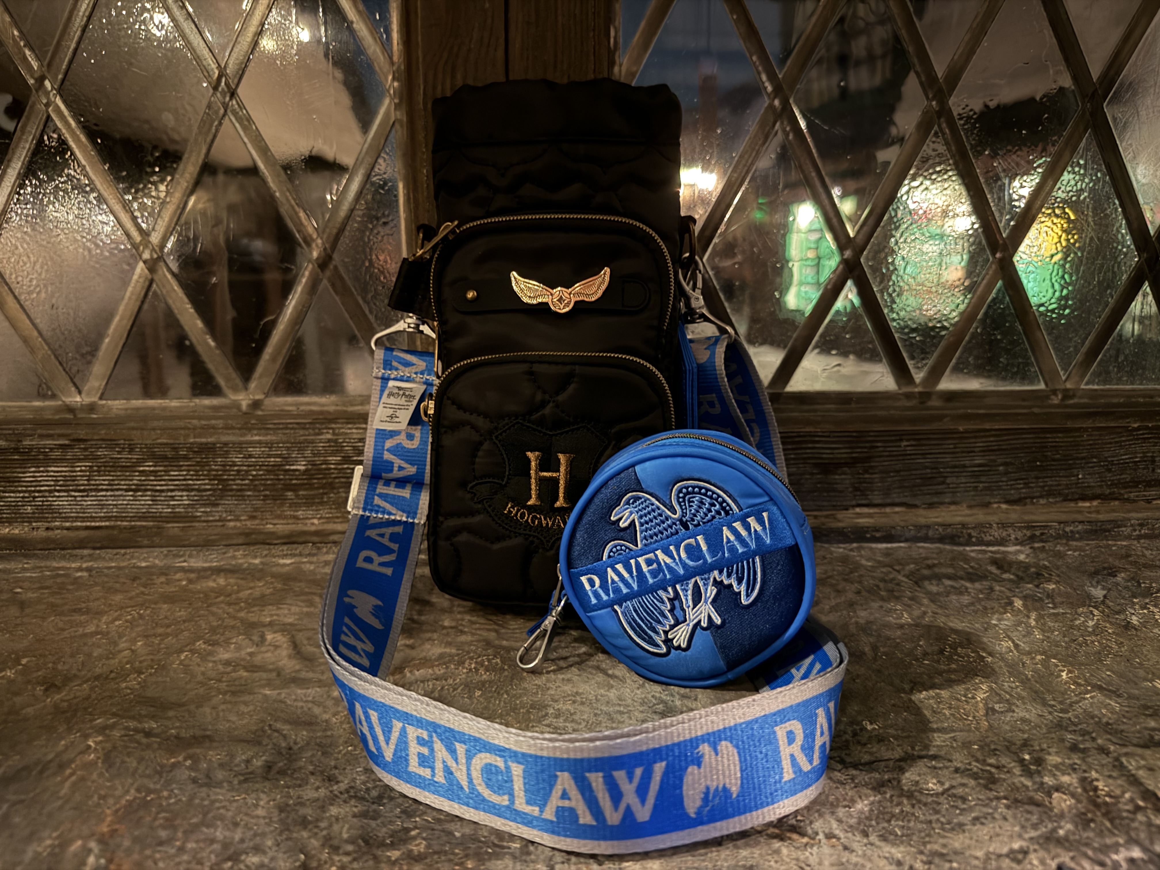 A black quilted Hogwarts Huggie water bottle bag with a gold Golden Snitch charm and embroidered crest, a round blue Ravenclaw coin pouch, and a blue and white RAVENCLAW strap coiled on a stone ledge at night