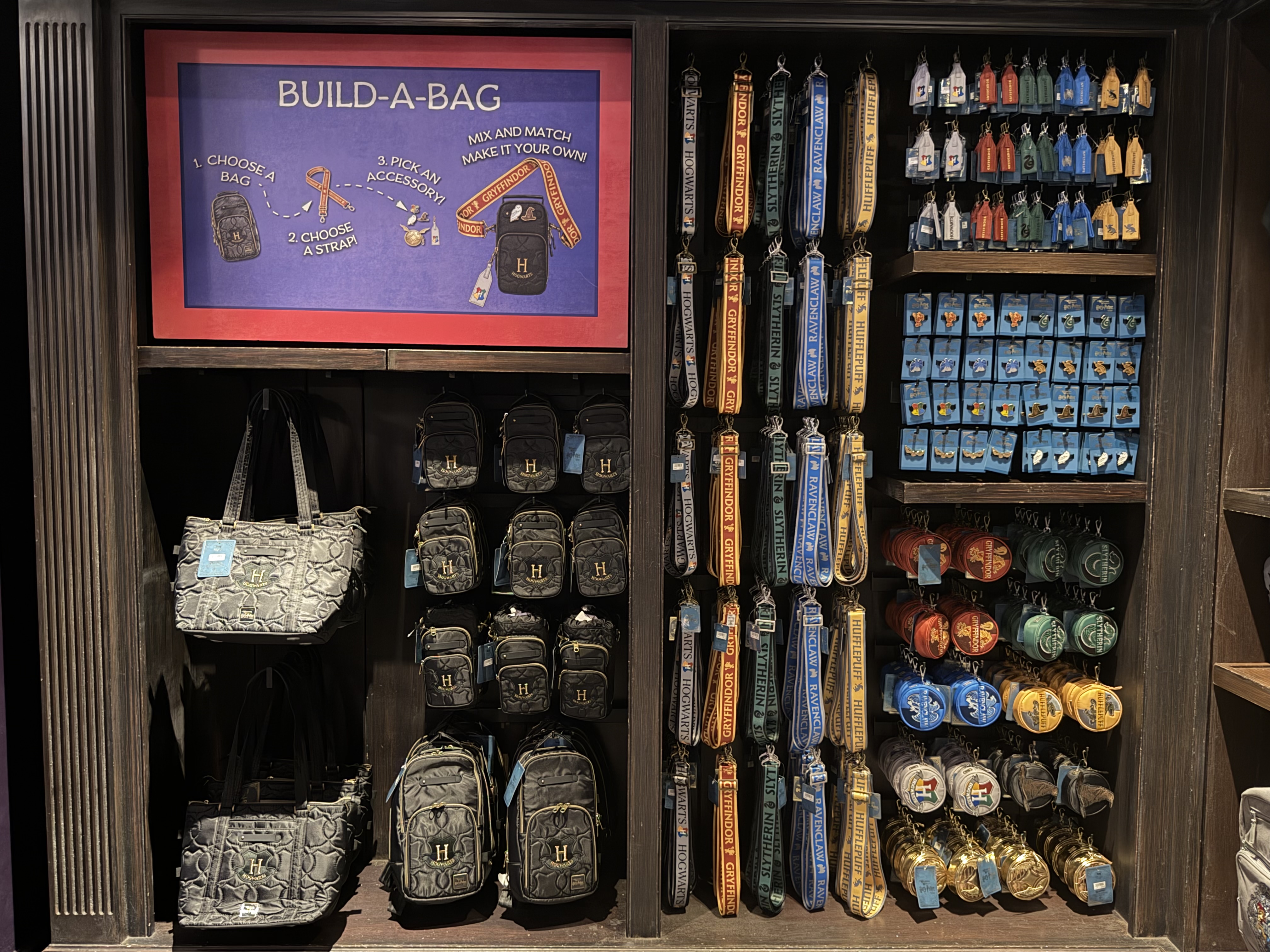 An in-park retail display with a lit BUILD-A-BAG sign showing three numbered steps, rows of black quilted Hogwarts bags, columns of house straps and pegs of charms and coin pouches