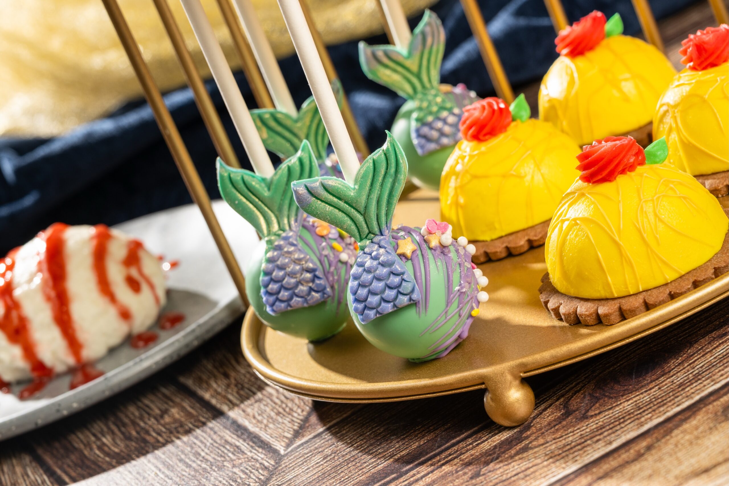 A gold pedestal stand holding green cake pops with purple scale collars and green mermaid tails, alongside yellow domed pastries decorated with red roses on cookie bases