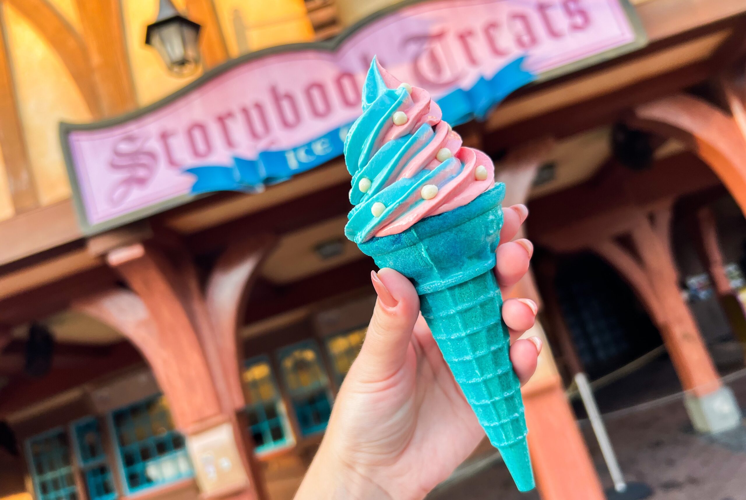 A hand holding a cone of pink and blue swirled soft-serve studded with small white pearls in a blue waffle-textured cone, in front of the Storybook Treats sign
