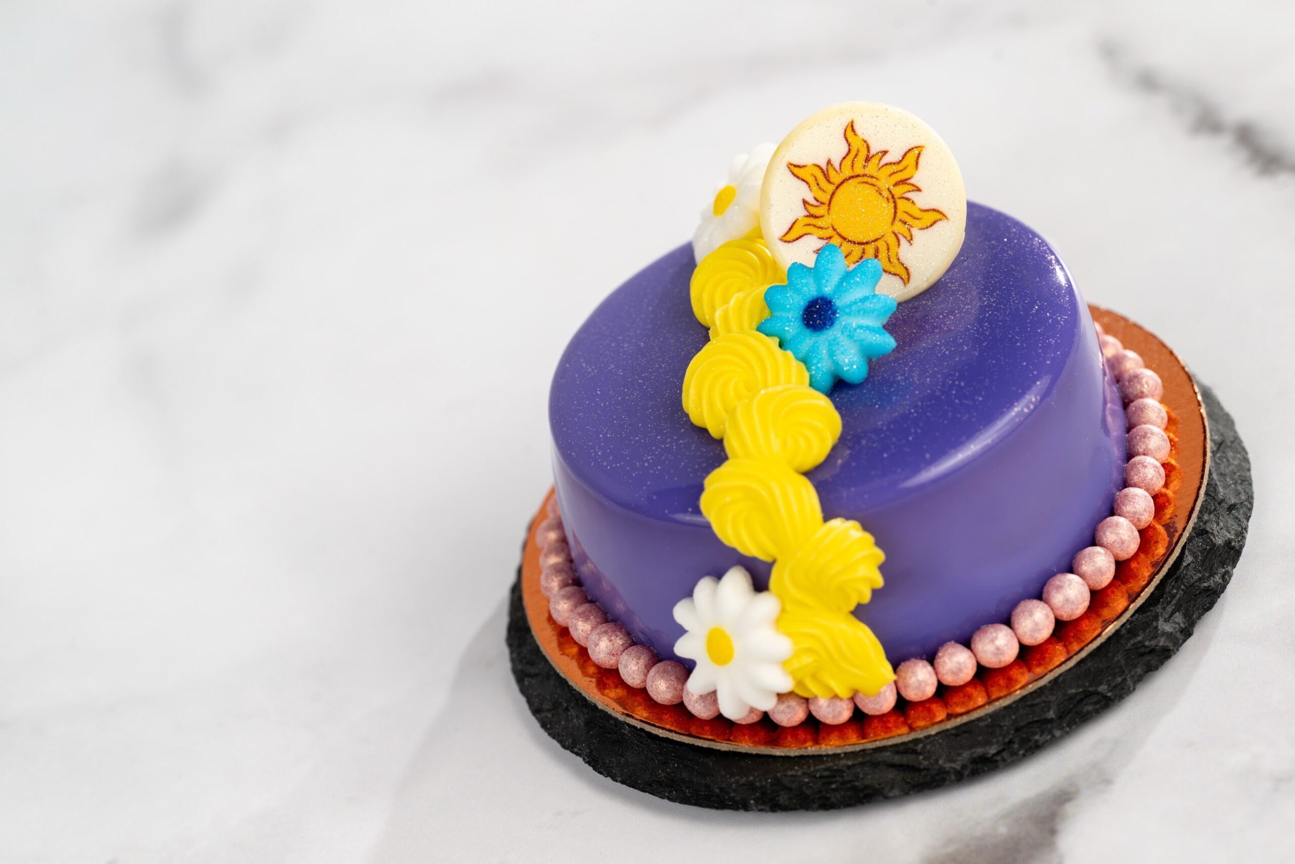 A round dessert with a glossy purple glaze, topped with a curving line of piped yellow frosting, white and blue sugar flowers, and a white chocolate disc printed with a golden sun emblem