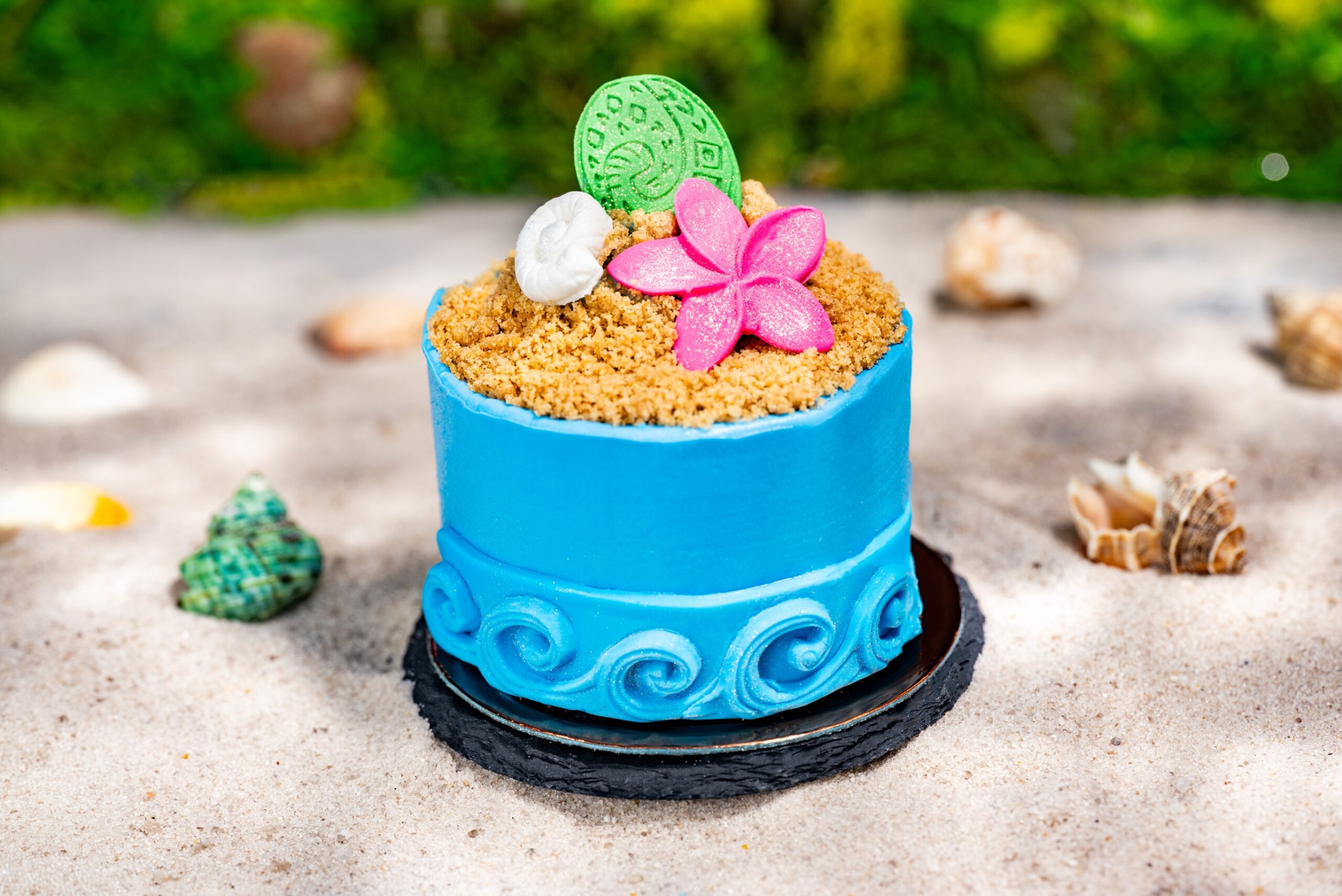 A small round cake covered in bright blue buttercream with sculpted wave detailing around the base, topped with dark chocolate and banana crumbs standing in for sand, a pink flower, a white shell and a green disc