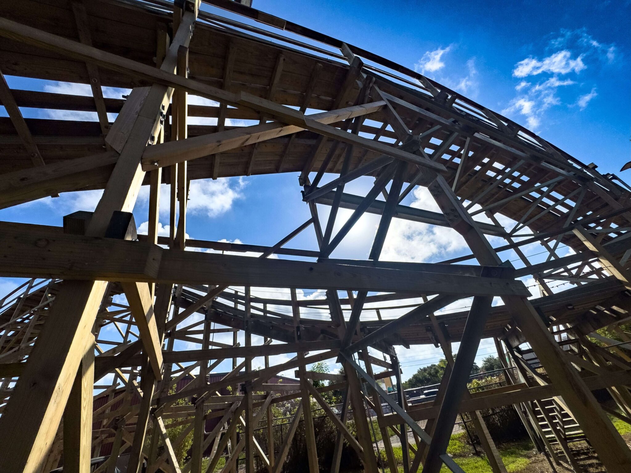 A view looking up through the timber framework of Coastersaurus, showing the banked curve of the track deck against a bright blue sky with scattered clouds.