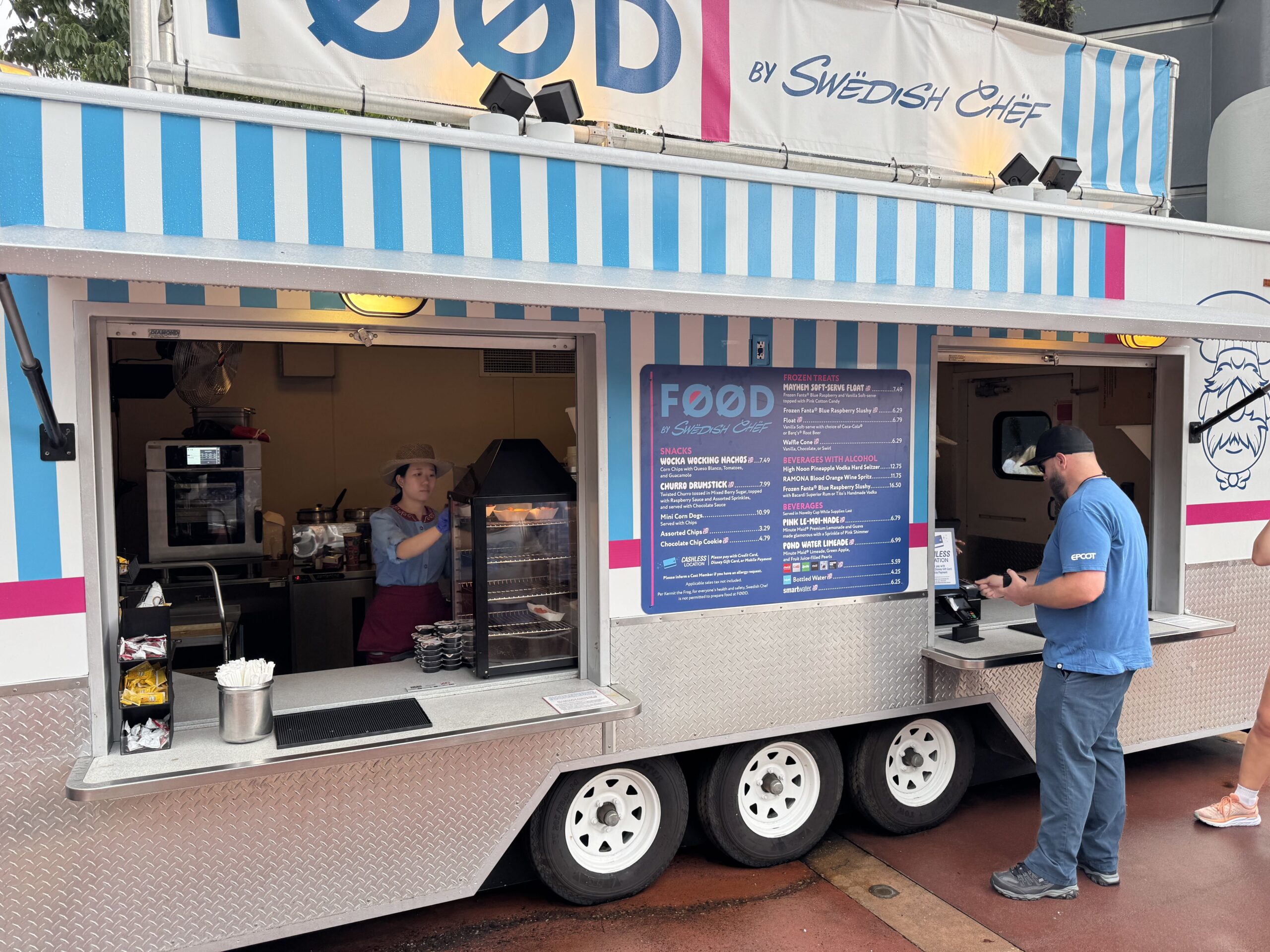 The FØØD by Swedish Chef kiosk, a blue, white and magenta striped trailer with a large menu board listing snacks, frozen treats and beverages, with a guest ordering at the window.