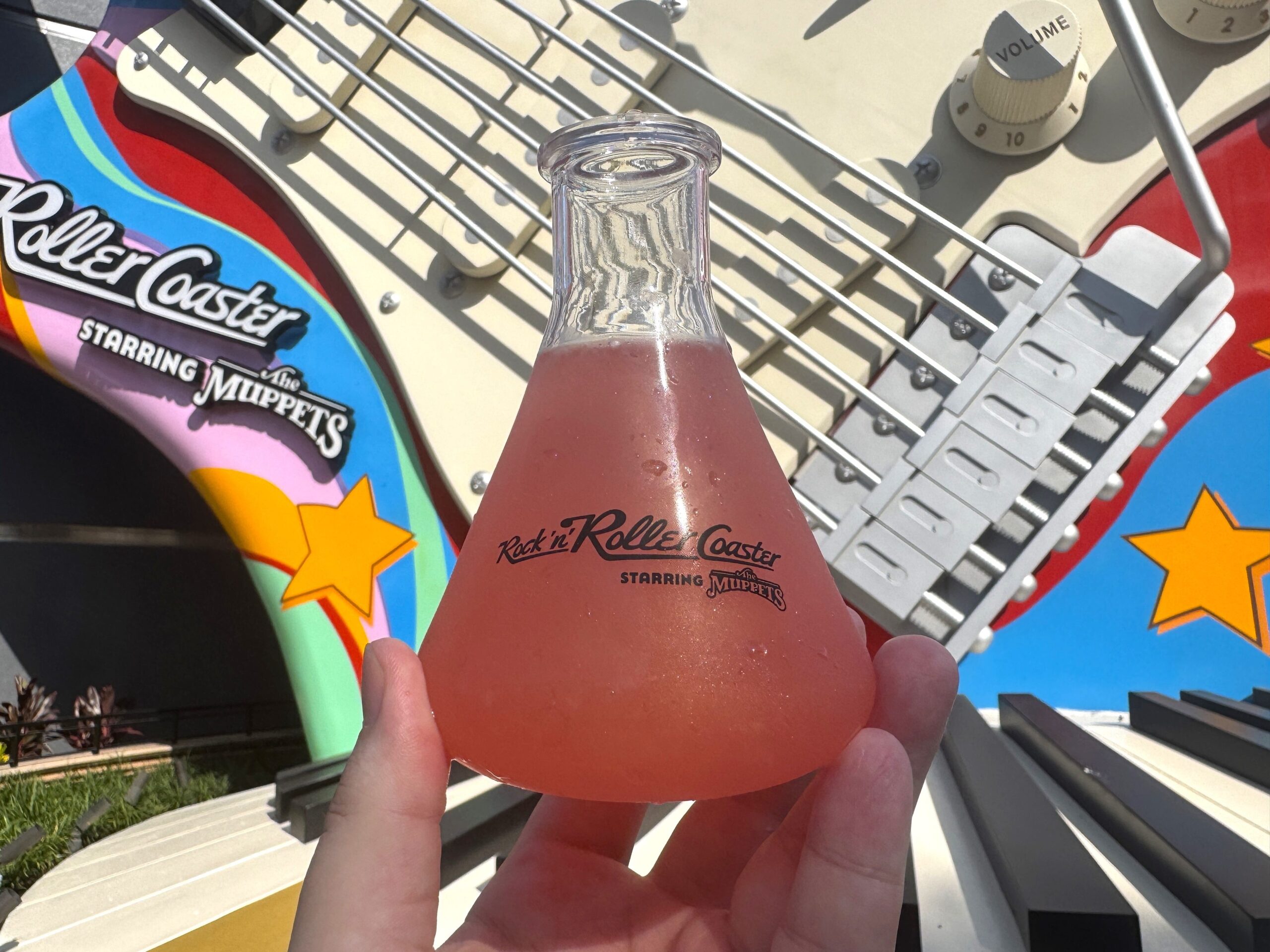 A clear conical flask filled with pink lemonade, printed with the Rock 'n' Roller Coaster Starring The Muppets wordmark, held up in front of the attraction's giant guitar and marquee.