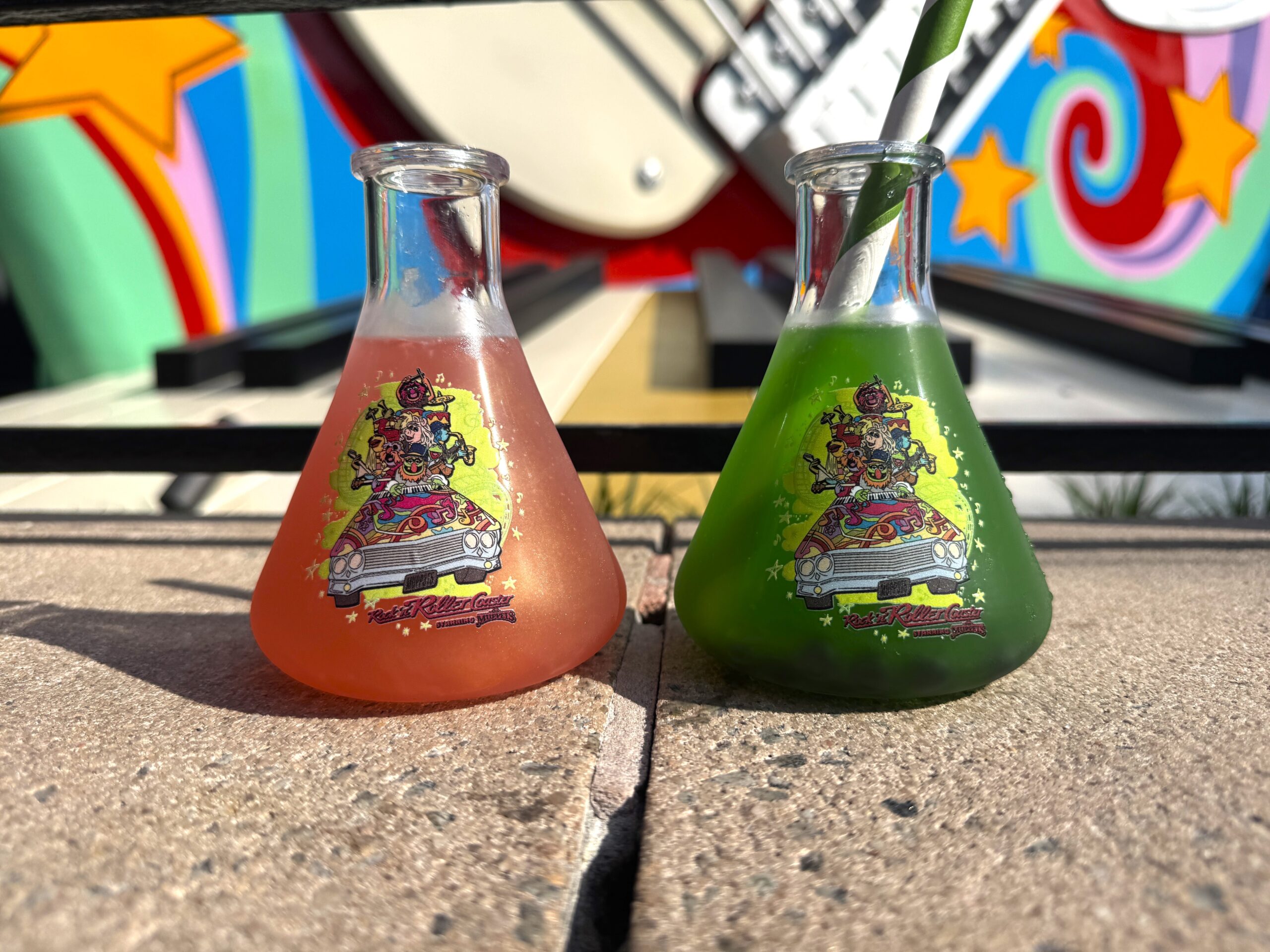 Two conical flask cups side by side on a ledge, one filled with pinkish orange lemonade and one with bright green limeade and a green striped straw, both printed with artwork of the Muppets crowded into a psychedelic limousine.