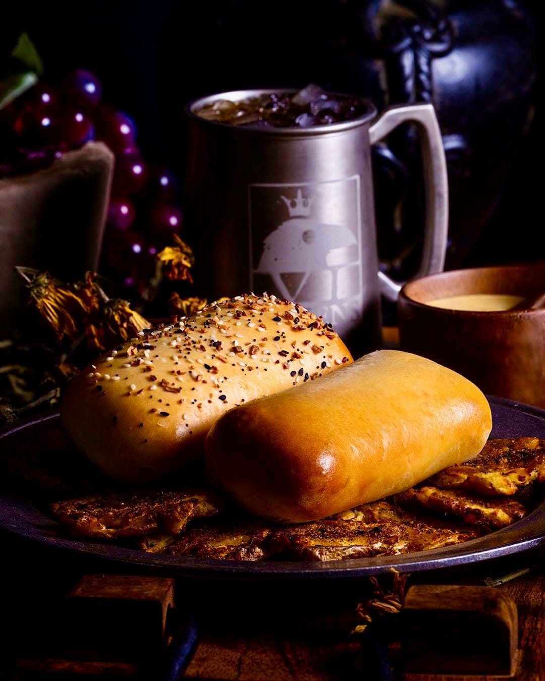 Two golden yeast roll Coffyns on a pewter plate, one topped with everything bagel seasoning and one plain, resting on seasoned smashed potatoes beside a wooden bowl of pale dipping sauce and a pewter stein bearing the crowned raven crest.