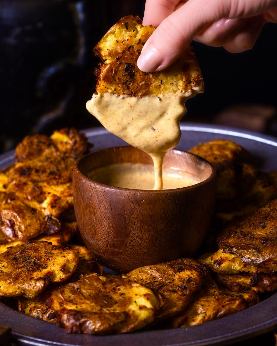 A hand lifts a crisp baked smashed potato dripping with pale yellow sauce out of a small wooden bowl, with more seasoned smashed potatoes arranged around it on a pewter plate.