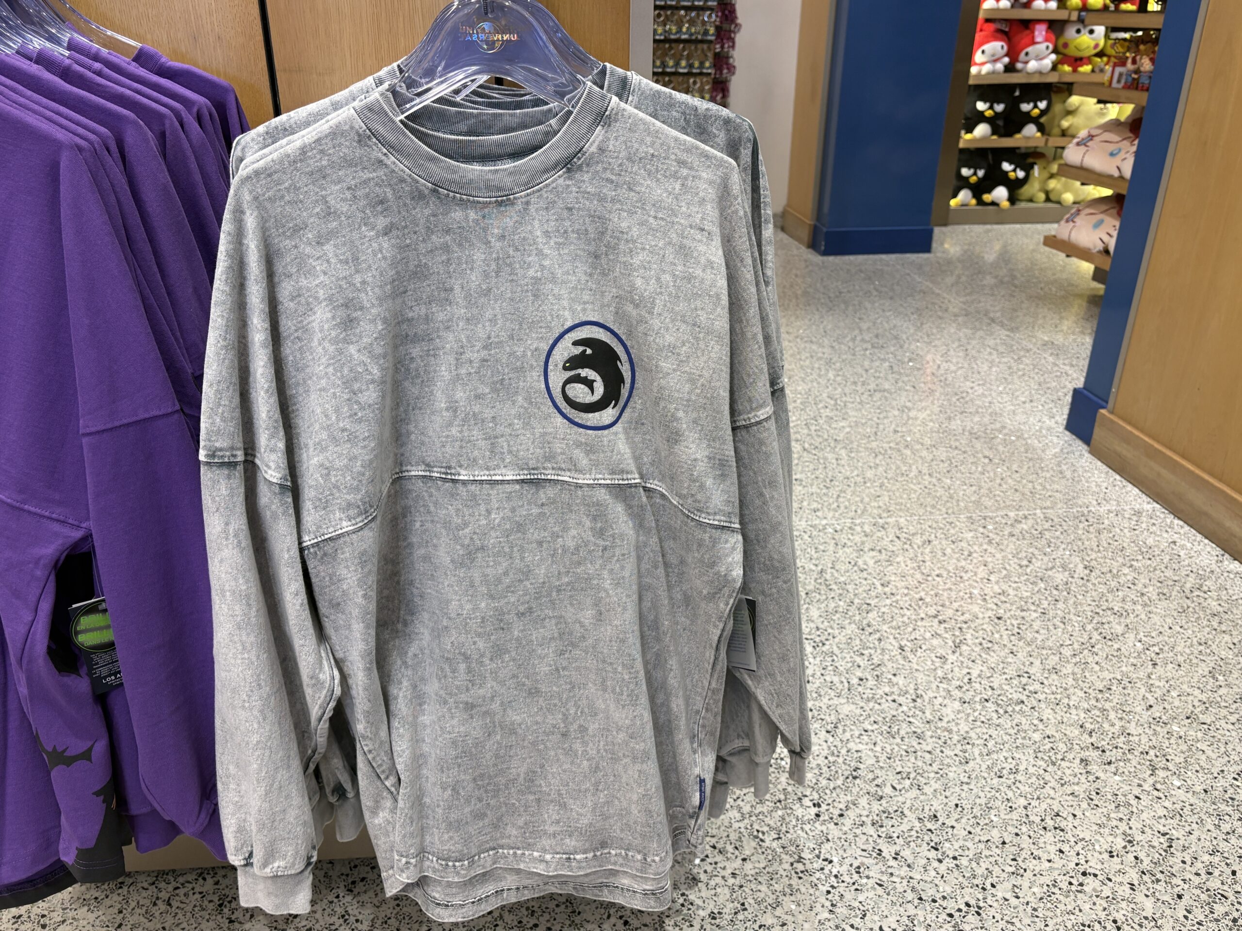 A gray acid wash Spirit Jersey on a hanger with the Night Fury emblem on the chest, a black dragon silhouette with one green eye curled inside a dark blue circle.
