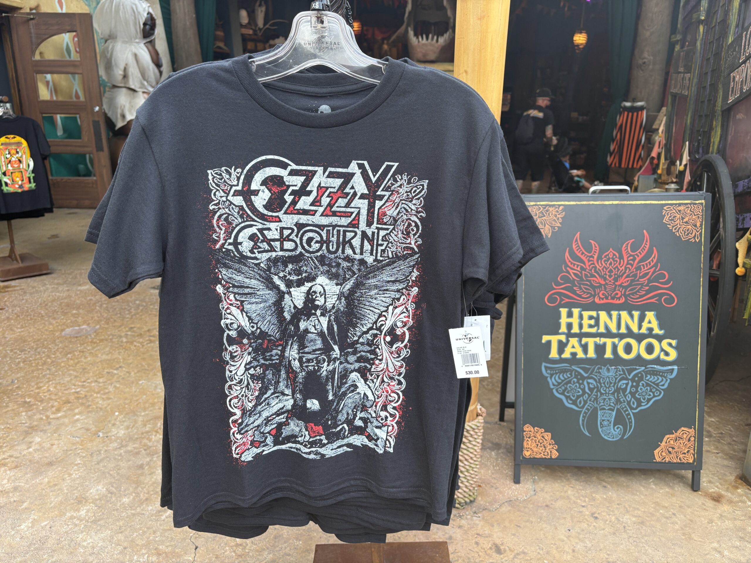 A black Ozzy Osbourne T-shirt on a hanger outside a themed storefront, printed with a stylized Ozzy Osbourne logo above a winged figure seated on a throne, with a Henna Tattoos sign to the right.
