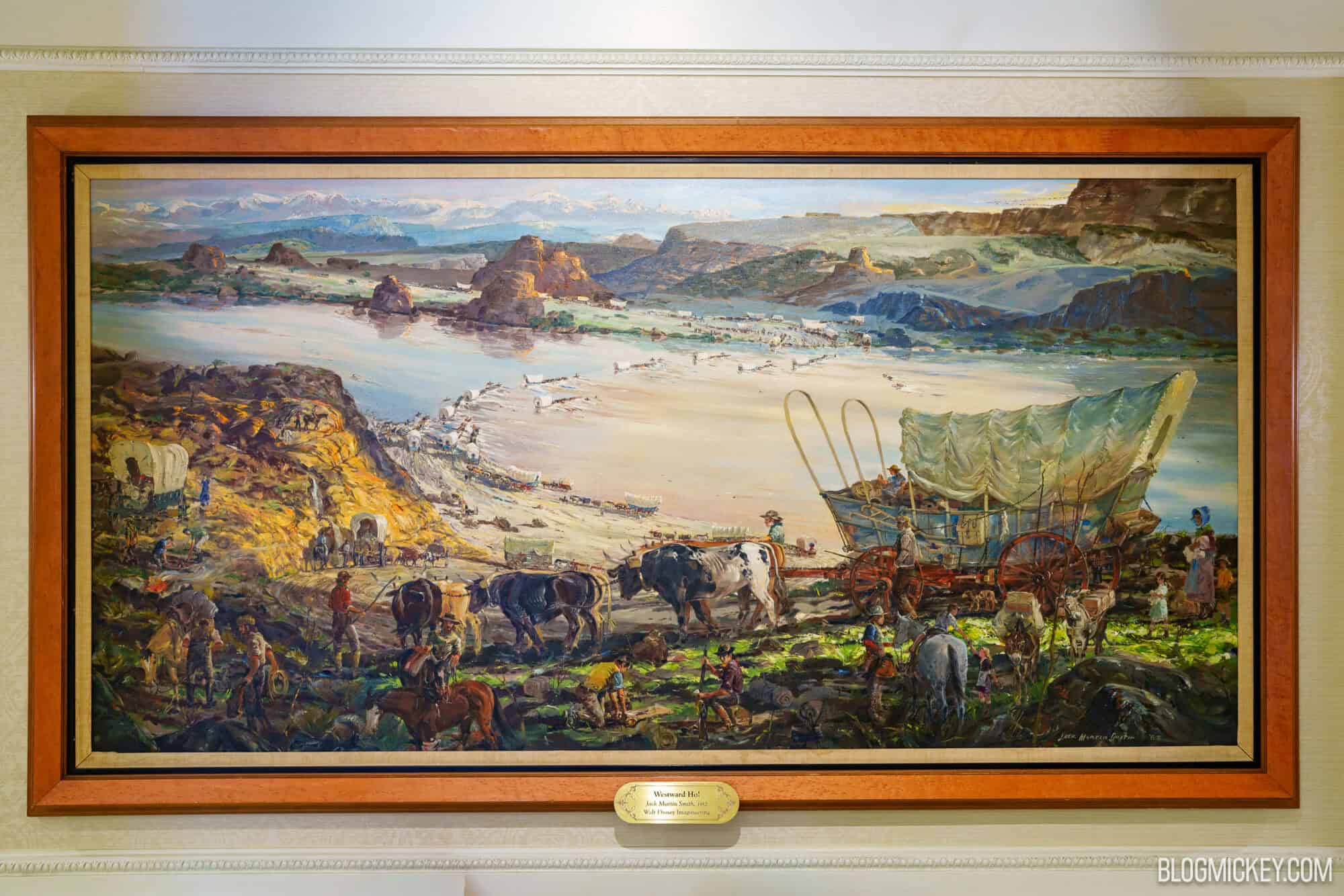 Westward Ho! by Jack Martin Smith, a panoramic painting with an ox team on a rocky trail in the foreground and a long line of covered wagons crossing a pale riverbed below snow-capped mountains