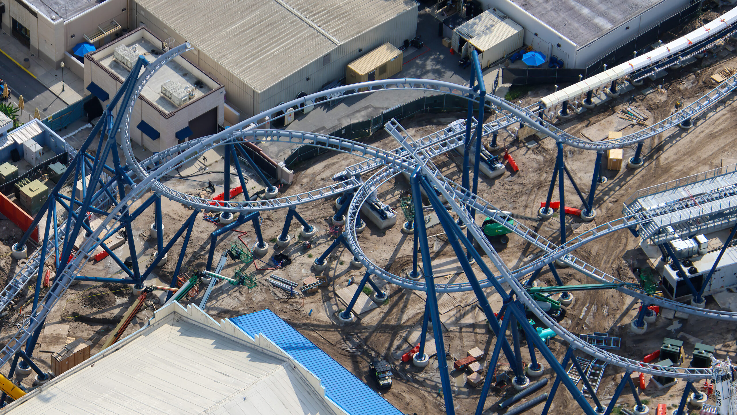 Daytime aerial photo of light gray coaster track crossing over and under itself above closely spaced dark blue support columns, with large park buildings along the edges of the frame, bare dirt below and construction equipment scattered across the site