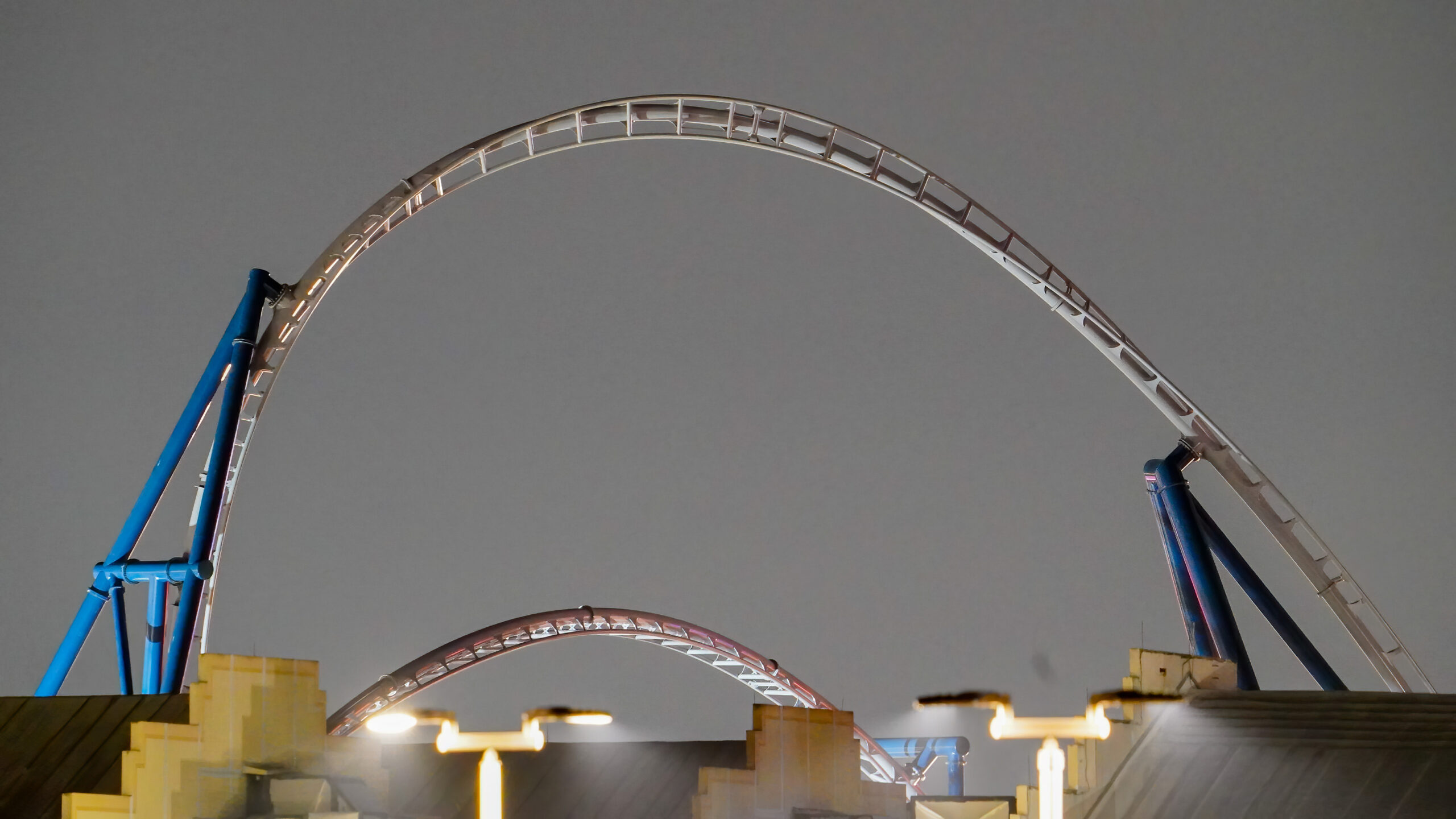 The completed upper arc of a roller coaster inversion, light gray track on dark blue supports, rising above soundstage rooftops against a flat gray pre-dawn sky, with a second lower arc of track behind it
