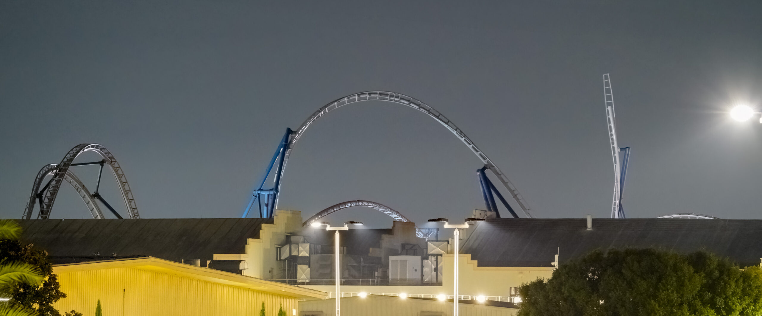 A wide pre-dawn view of several roller coaster track elements, including a tall arch on blue supports at center and an angled spike element at right, rising above a long dark soundstage roofline, with a yellow lit warehouse building and tall light poles in the foreground