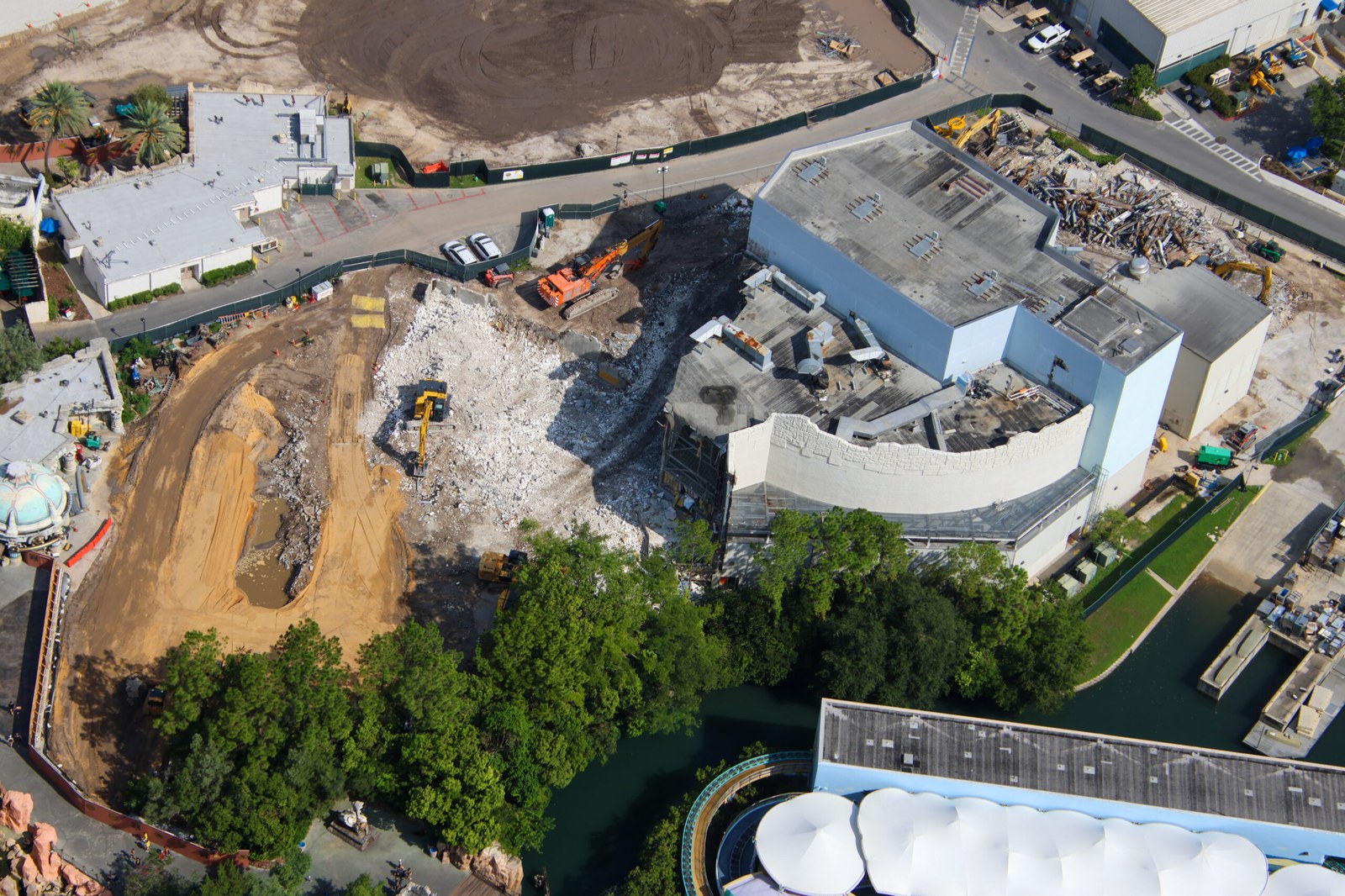 Aerial view of a wide field of broken white concrete debris with orange and yellow excavators working around it, beside a partially demolished show building with a curved white themed wall still standing.