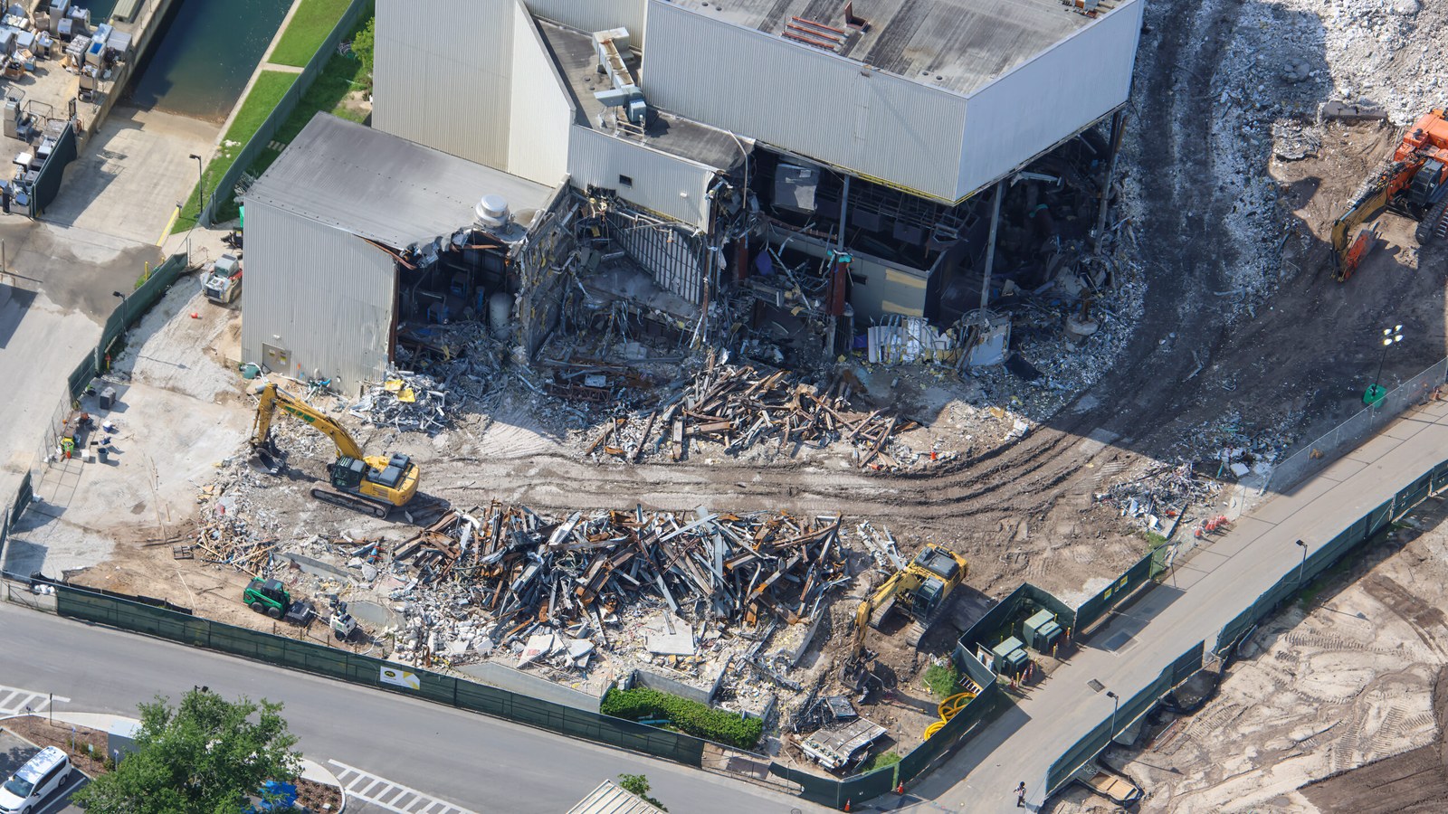 Aerial view of long piles of rusted steel beams and construction debris beside a partially demolished building, with two yellow excavators and green construction walls along a backstage road.