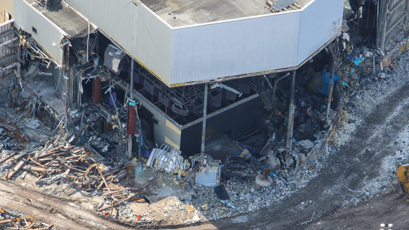 Close aerial view of a corner of the Poseidon's Fury show building with its exterior walls torn away, exposing bare structural columns and dark interior walls, with piles of rusted steel beams on the ground below.
