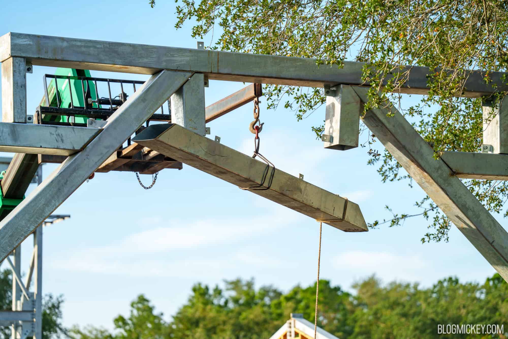 Close view of galvanized steel framing against a blue sky, with a single steel beam hanging from a crane hook and sling, a yellow tag line dangling from its end, and a green lift at left