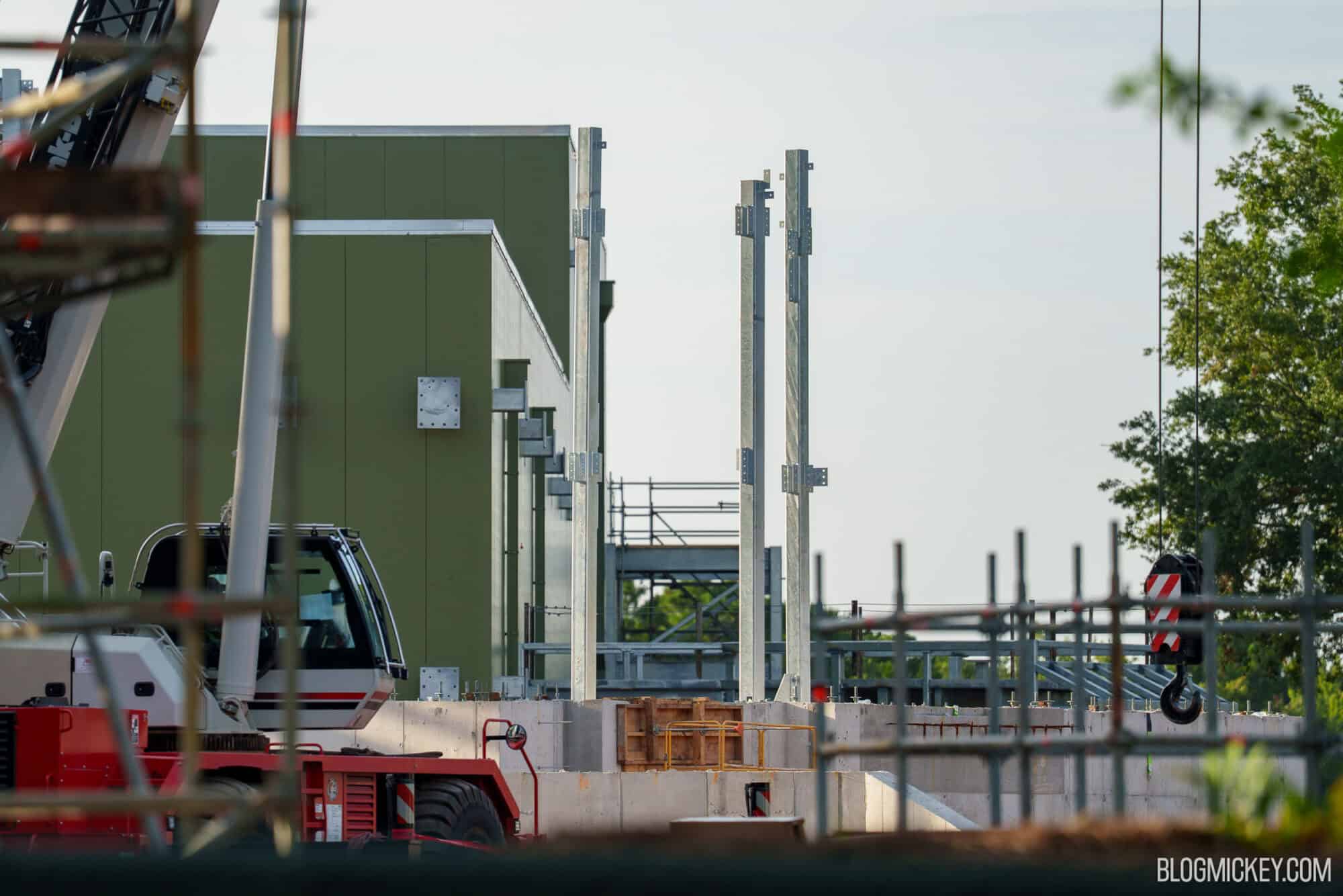 Three bare galvanized steel columns with bolt plates standing on a poured concrete wall with a large olive green building behind them, with a red and white mobile crane in the foreground and a crane hook hanging at right