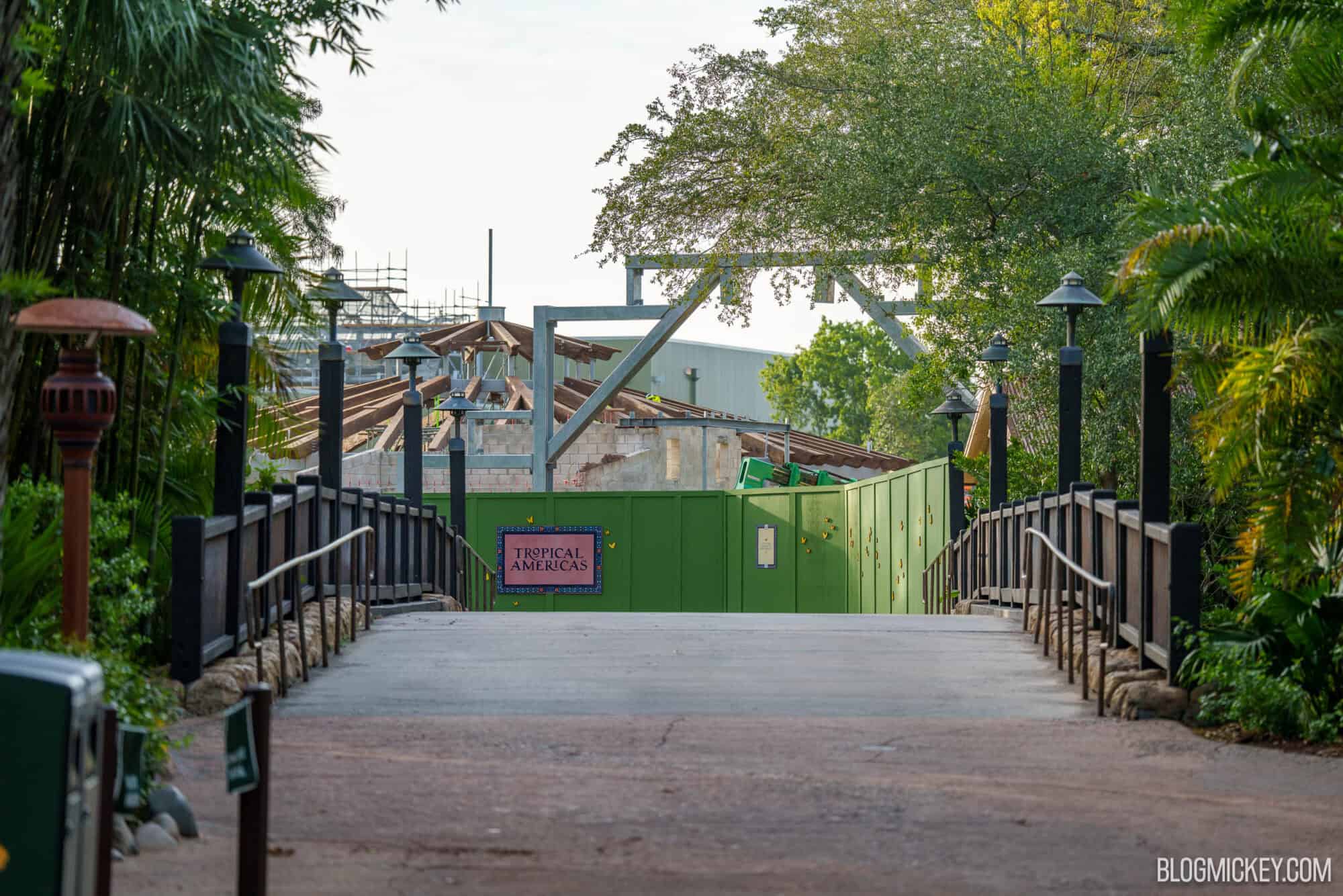 View up a paved walkway and over a wooden bridge toward a green construction wall bearing a Tropical Americas sign, with galvanized steel beams, blond wooden roof rafters and a pale green show building rising behind it