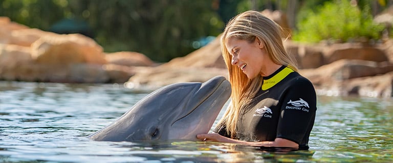 A guest in a Discovery Cove wetsuit stands chest deep in the lagoon beside a bottlenose dolphin surfacing next to her