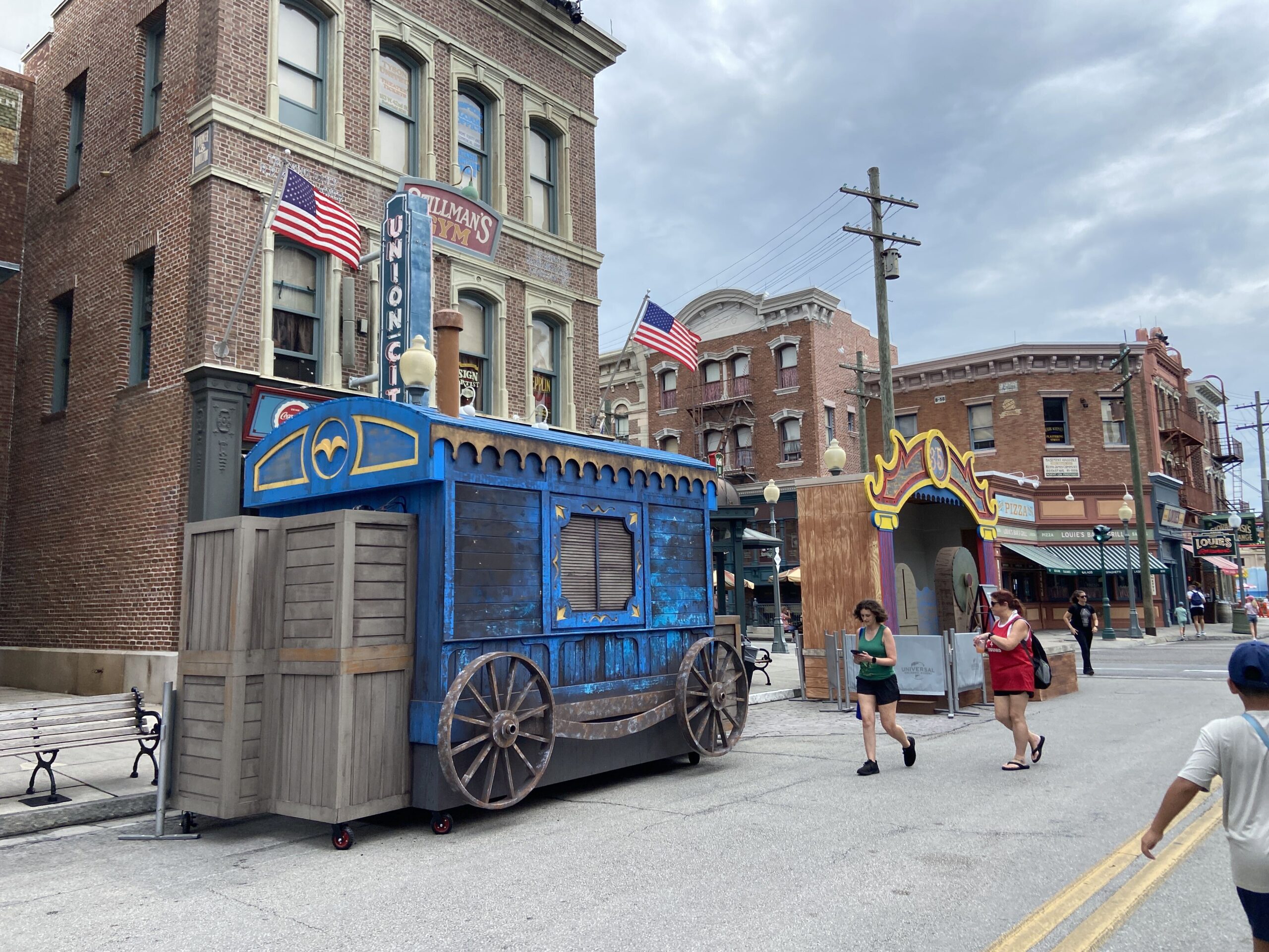 A blue and gold carnival wagon set piece with large spoked wheels and stacked wooden crates, parked on the New York backlot street