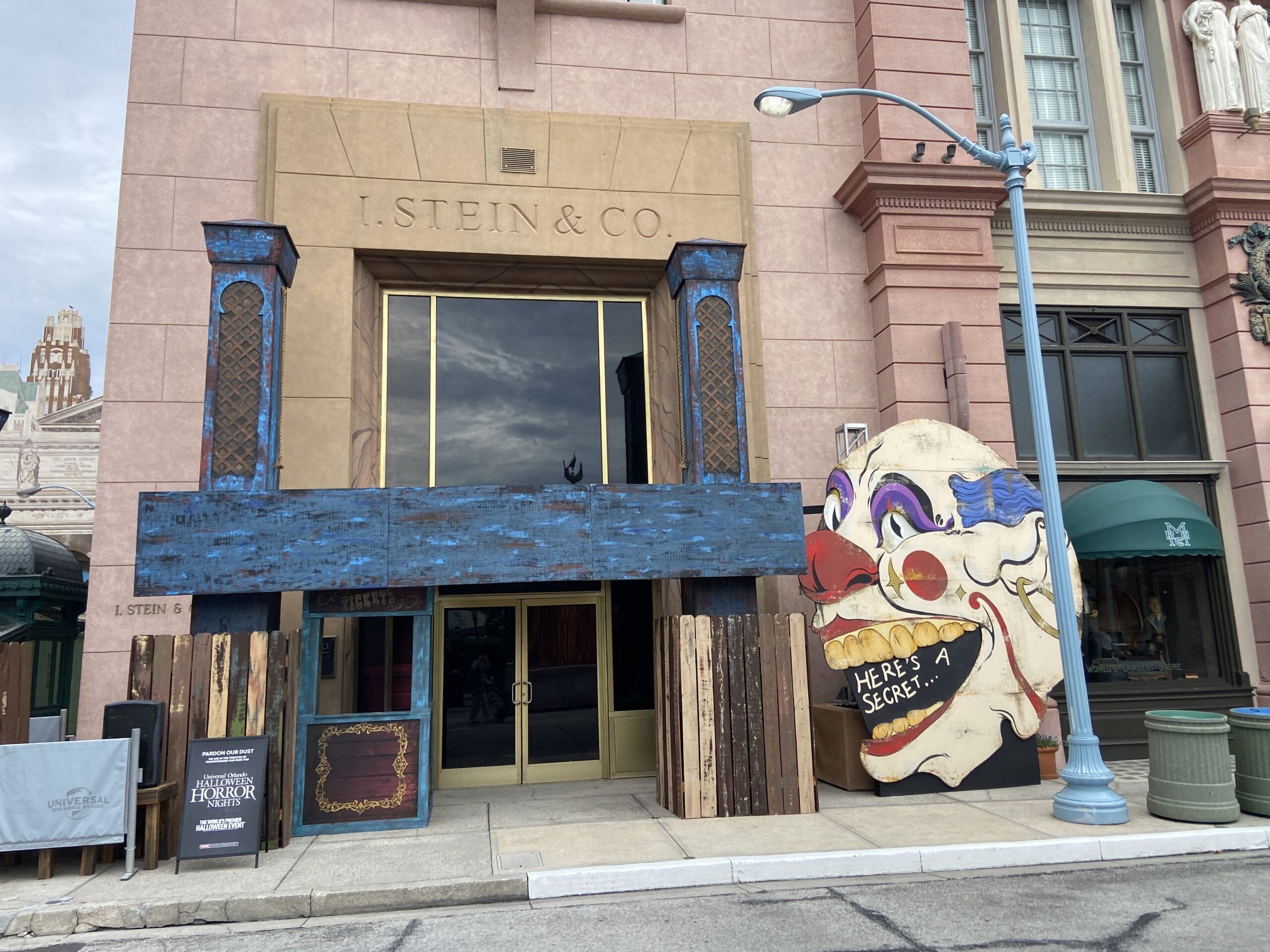 The I. Stein and Co. facade housing the Tribute Store, flanked by weathered wooden barricade fencing under a blue painted canopy board, with a large painted clown face cutout standing at the right side