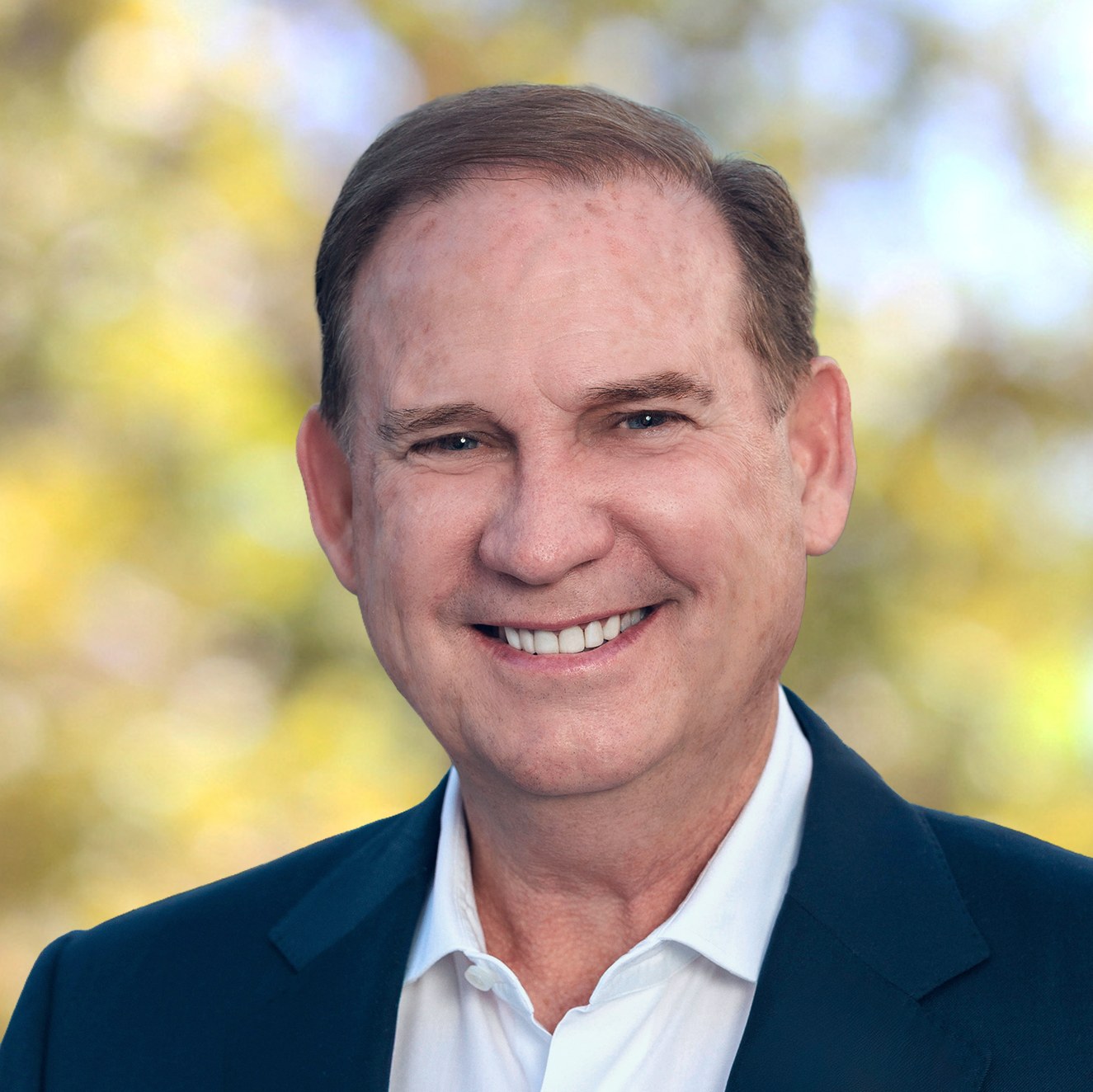 Corporate portrait of Joe Schott in a navy blazer and white shirt, smiling, against a blurred green and yellow background