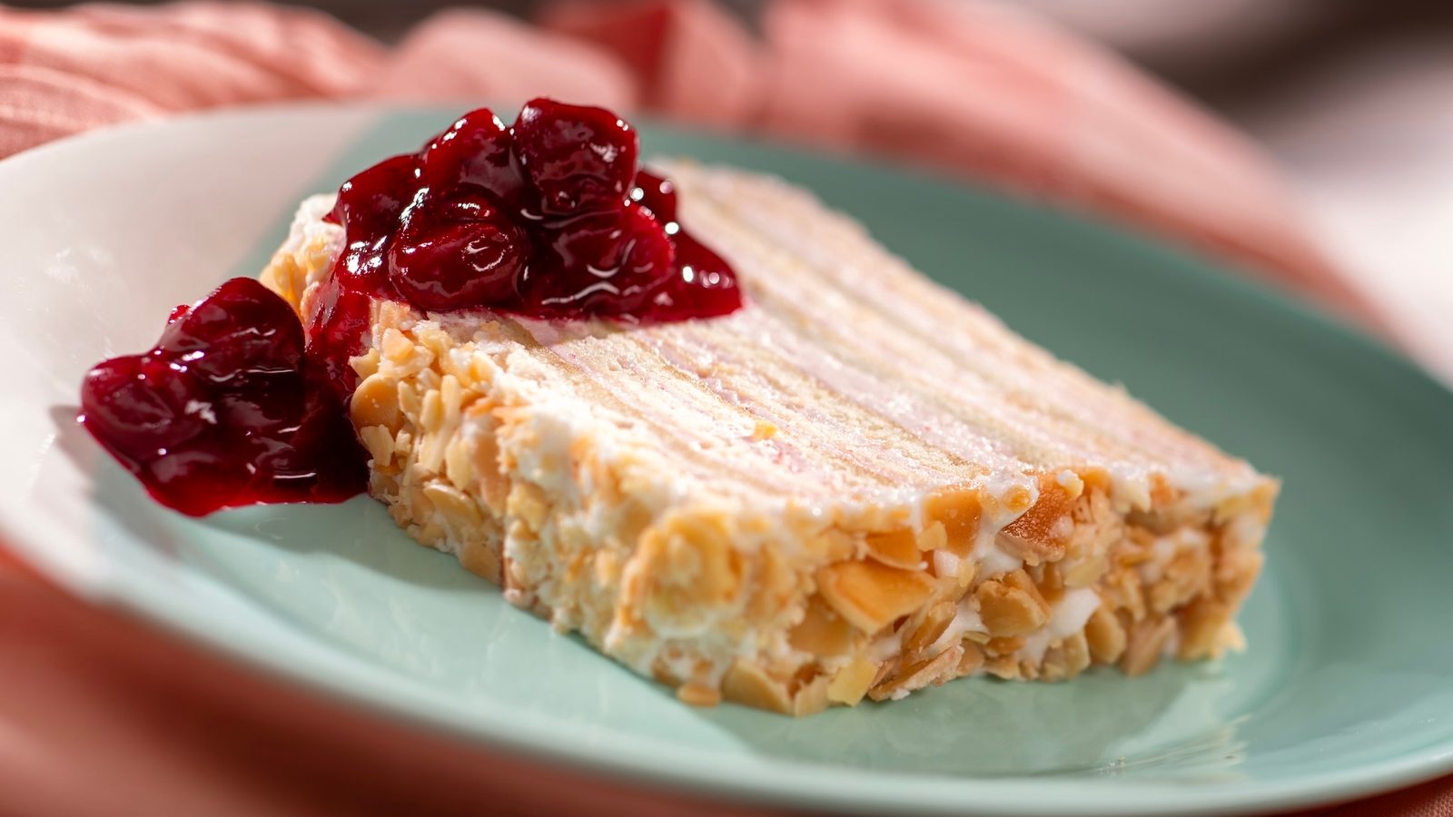 A slice of layered Kirschwasser Torte on a pale green plate, edged in sliced almonds and topped with dark cherry compote