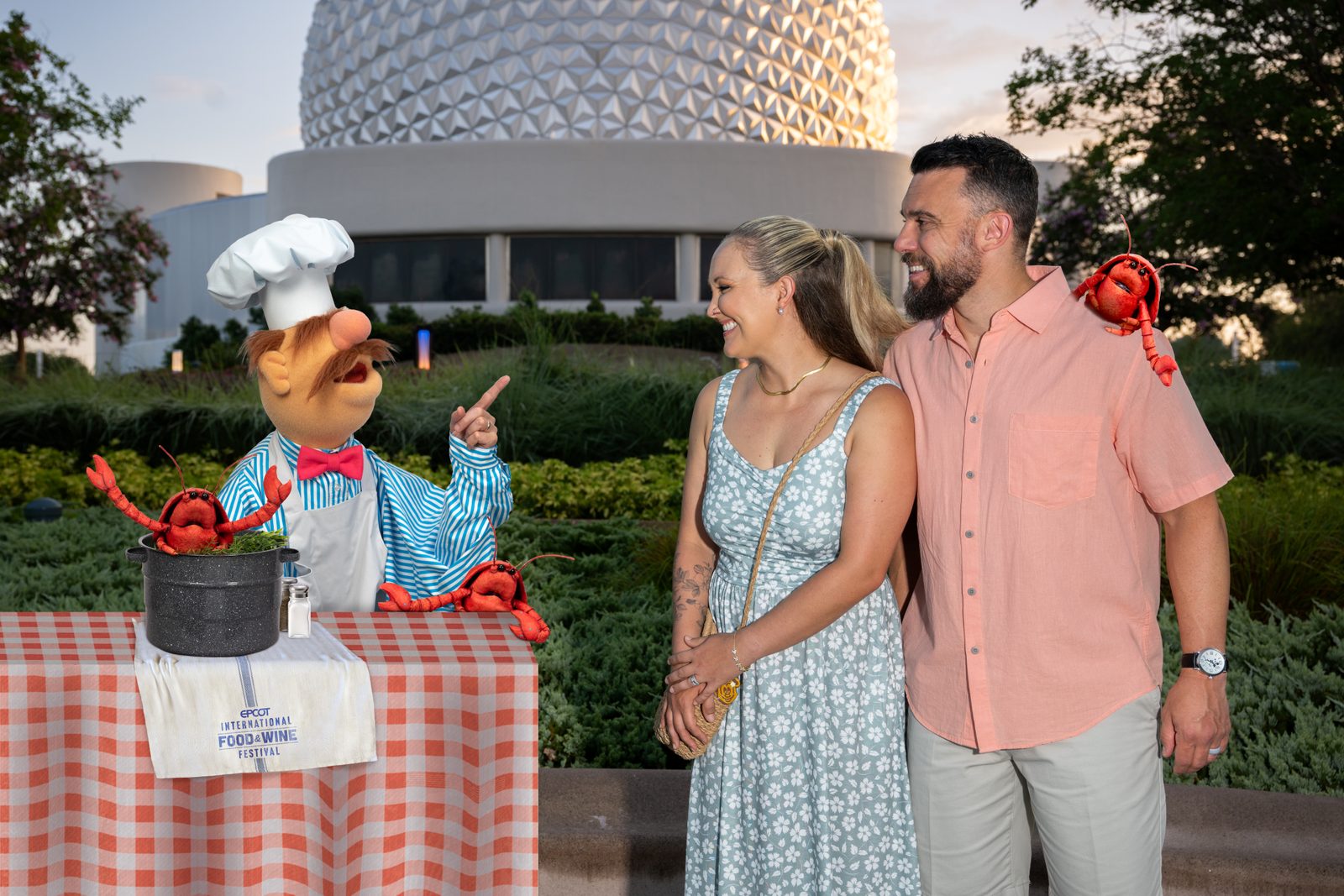 The Swedish Chef stands behind a checkered table with a stockpot of plush lobsters and an EPCOT International Food and Wine Festival towel, pointing at a couple with a lobster perched on one shoulder, Spaceship Earth behind them