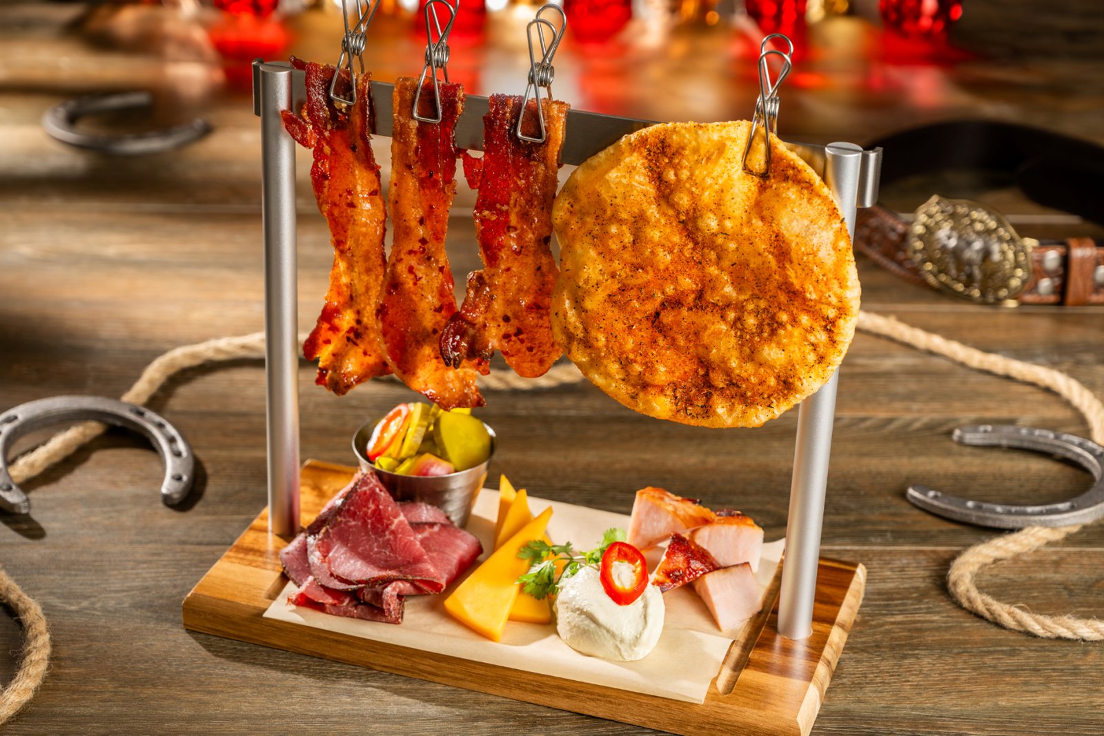 Close view of the charcuterie board, with bacon strips and a large crisped cheese round clipped to a metal stand above sliced meats, cheddar wedges and pickles