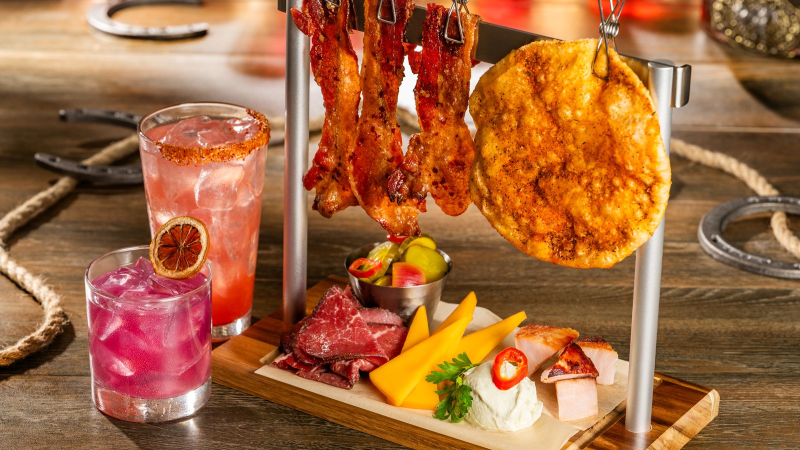 A charcuterie board with bacon hanging from a metal rack, next to a pink margarita and a chili rimmed lemonade on a wooden saloon table