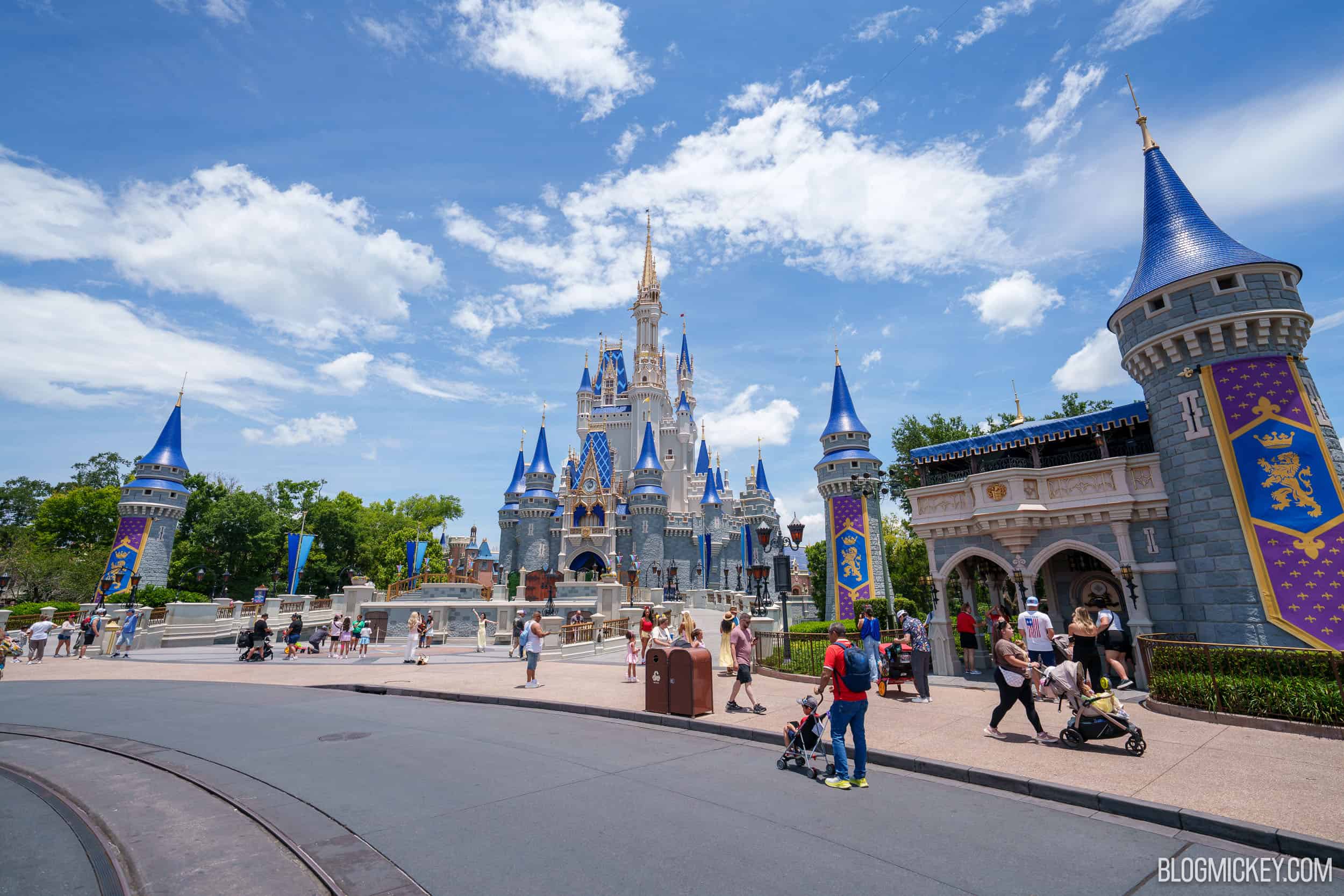 A wide daytime view of Cinderella Castle from the Magic Kingdom hub, with a scattering of guests and strollers walking the pavement and heraldic banners hanging at right.