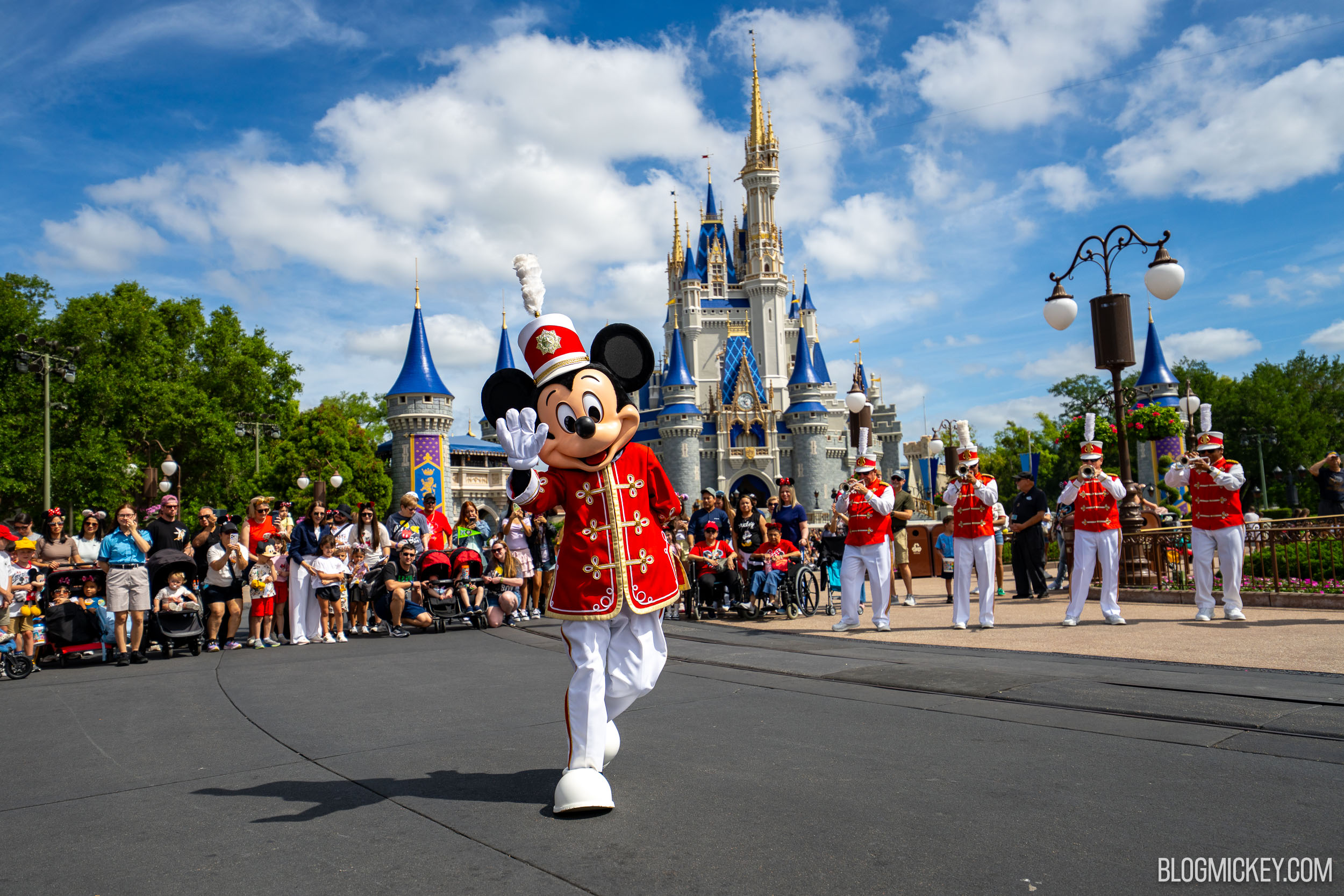 Mickey Mouse in a red and gold bandleader coat waves while marching across open pavement in the Magic Kingdom hub, ahead of brass players in matching red coats and plumed shakos, with guests lined one deep along the route and Cinderella Castle behind him.