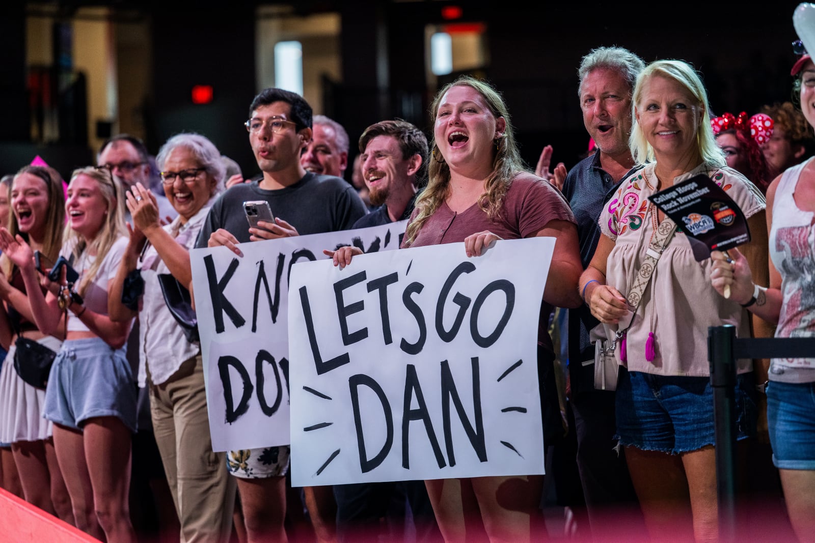 A crowd of spectators cheers and claps behind a barrier, two of them holding hand lettered signs reading LET'S GO DAN