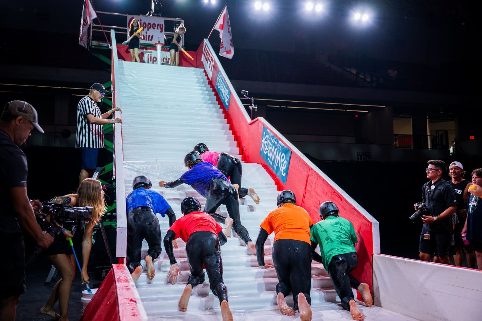 Competitors in colored shirts and helmets crawl up a soaked white staircase while a referee watches and camera operators film from the side
