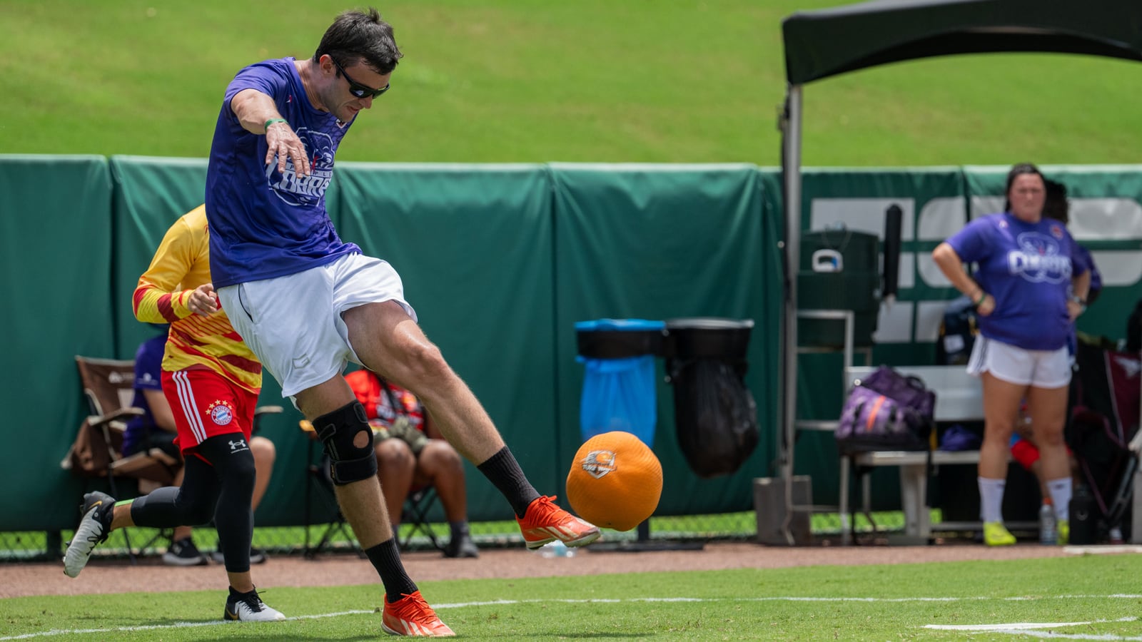 A man in a purple shirt and white shorts kicks a large orange rubber ball on a grass field as teammates watch from the sideline