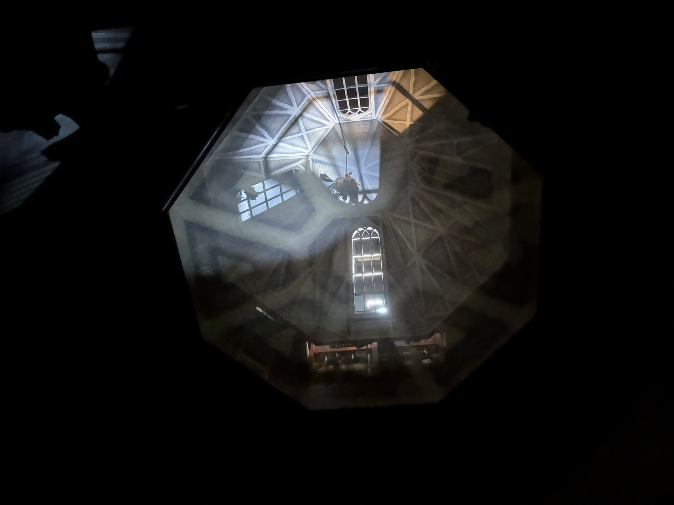 Looking up through the new scrim into the dark attic space above, where a figure hangs from a rope in the upper third of the lit octagon beside painted arched windows and rafters