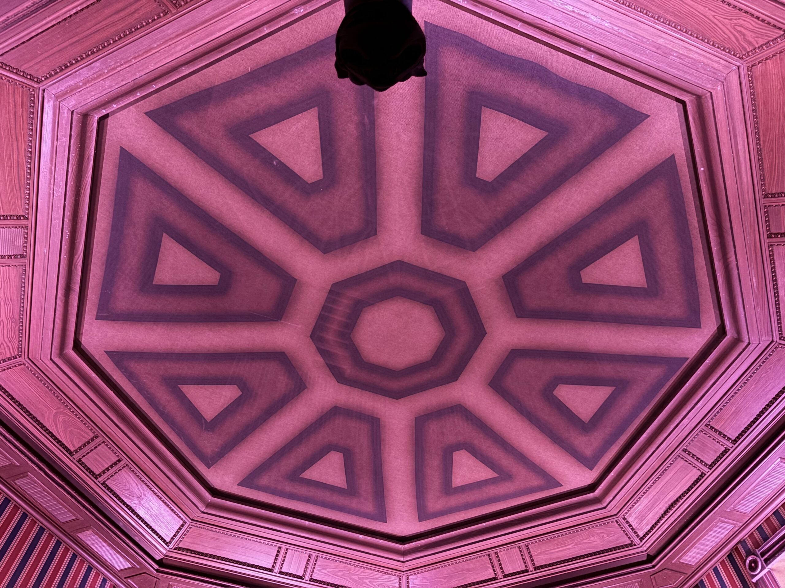 The octagonal Stretching Room ceiling at Magic Kingdom lit in bright pink, showing the new scrim with a radiating pattern of triangles around a center octagon